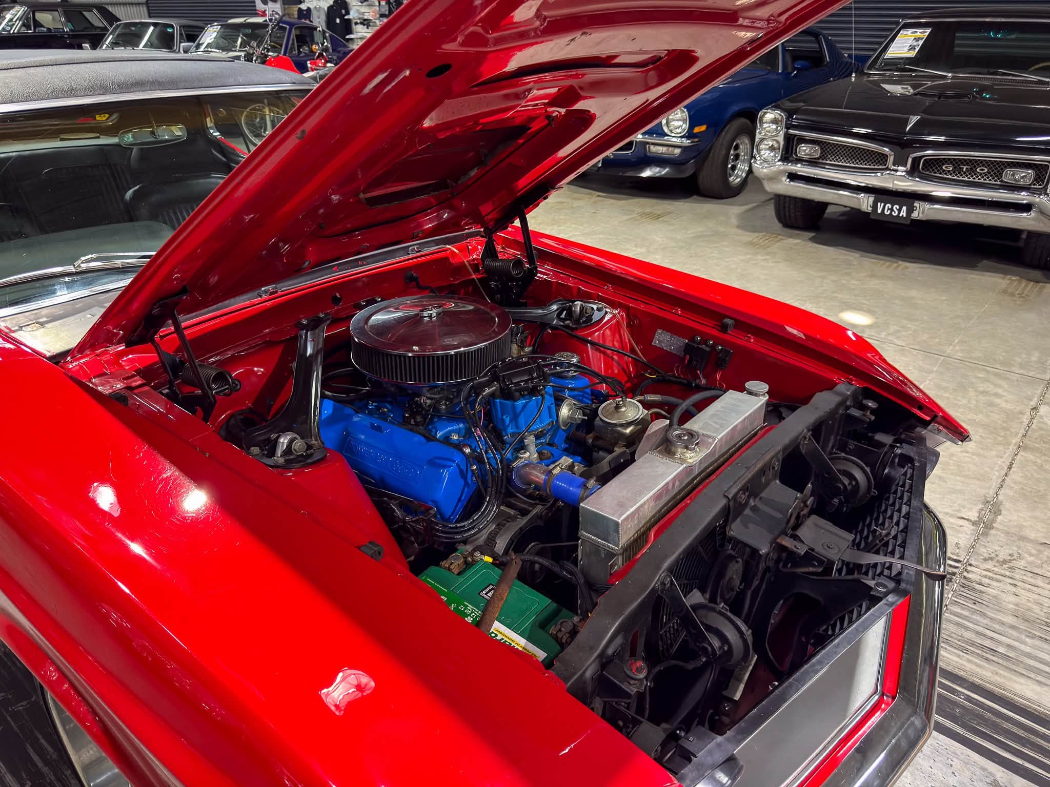 Red car with hood open, showing a blue engine block in a classic car on display in a showroom.