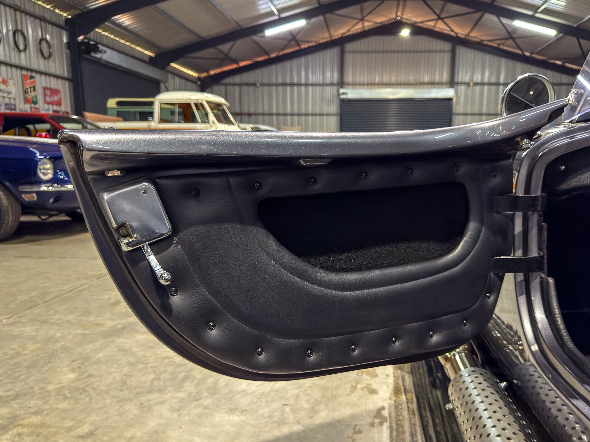 Open car door interior of a vintage black convertible, with chrome trim and leather panels, in a garage with other classic cars in the background.