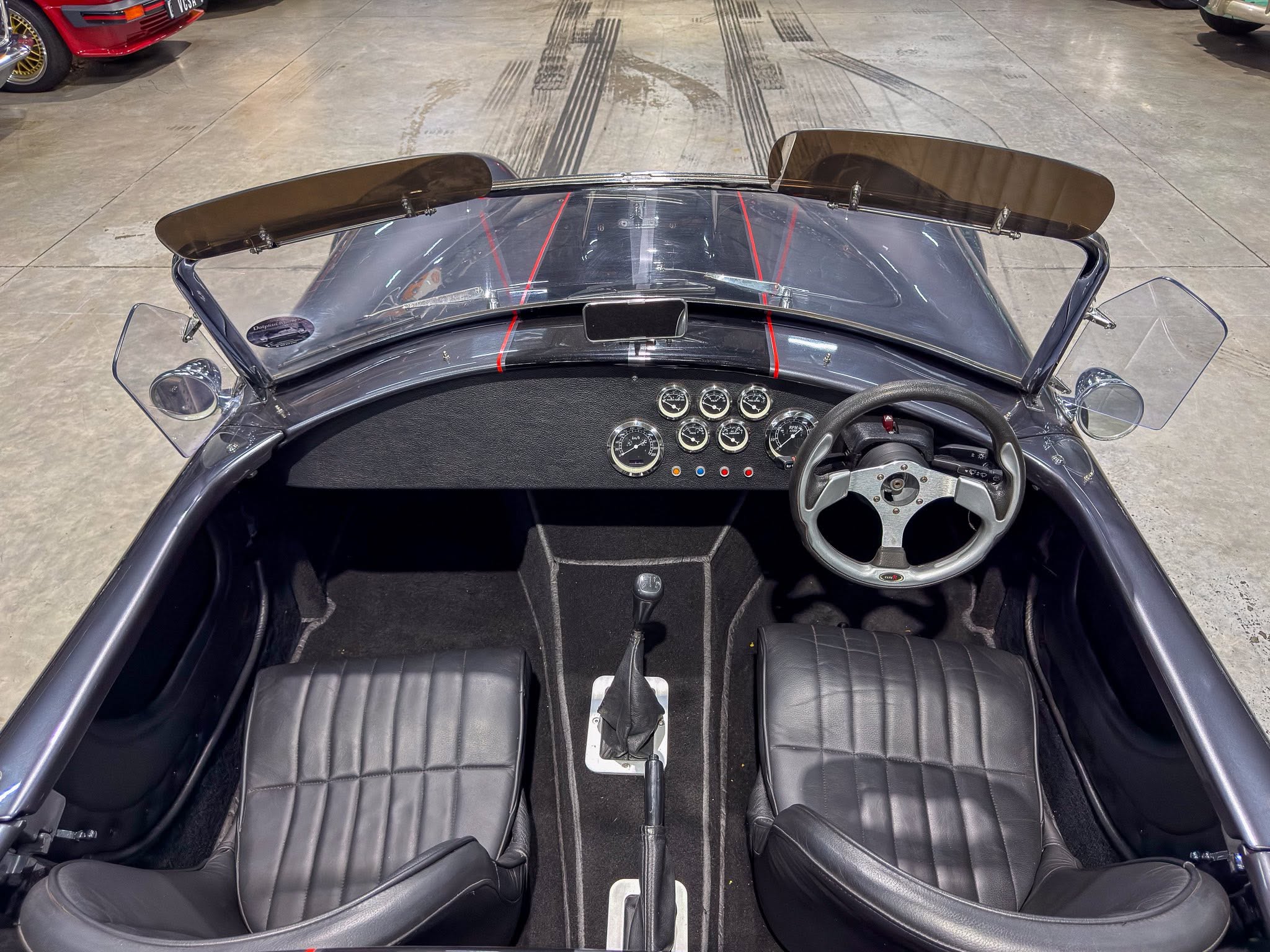 From the driver’s seat of a classic convertible, black leather seats, analog gauges, and a manual shift lever, with pop-up wind deflectors and a gray dashboard in a showroom.