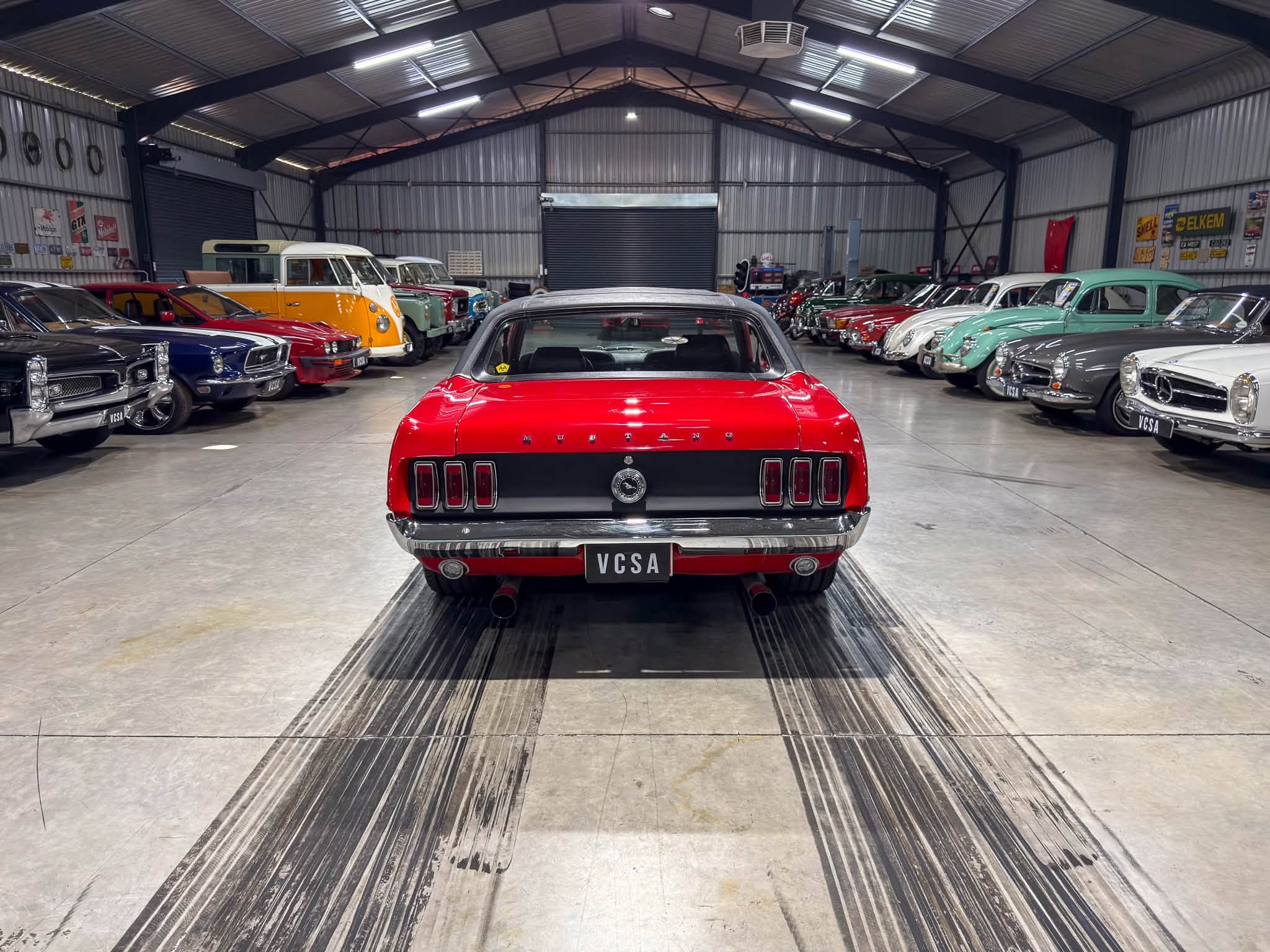 Rear view of a red Ford Mustang in a spacious showroom lined with rows of vintage cars on both sides.