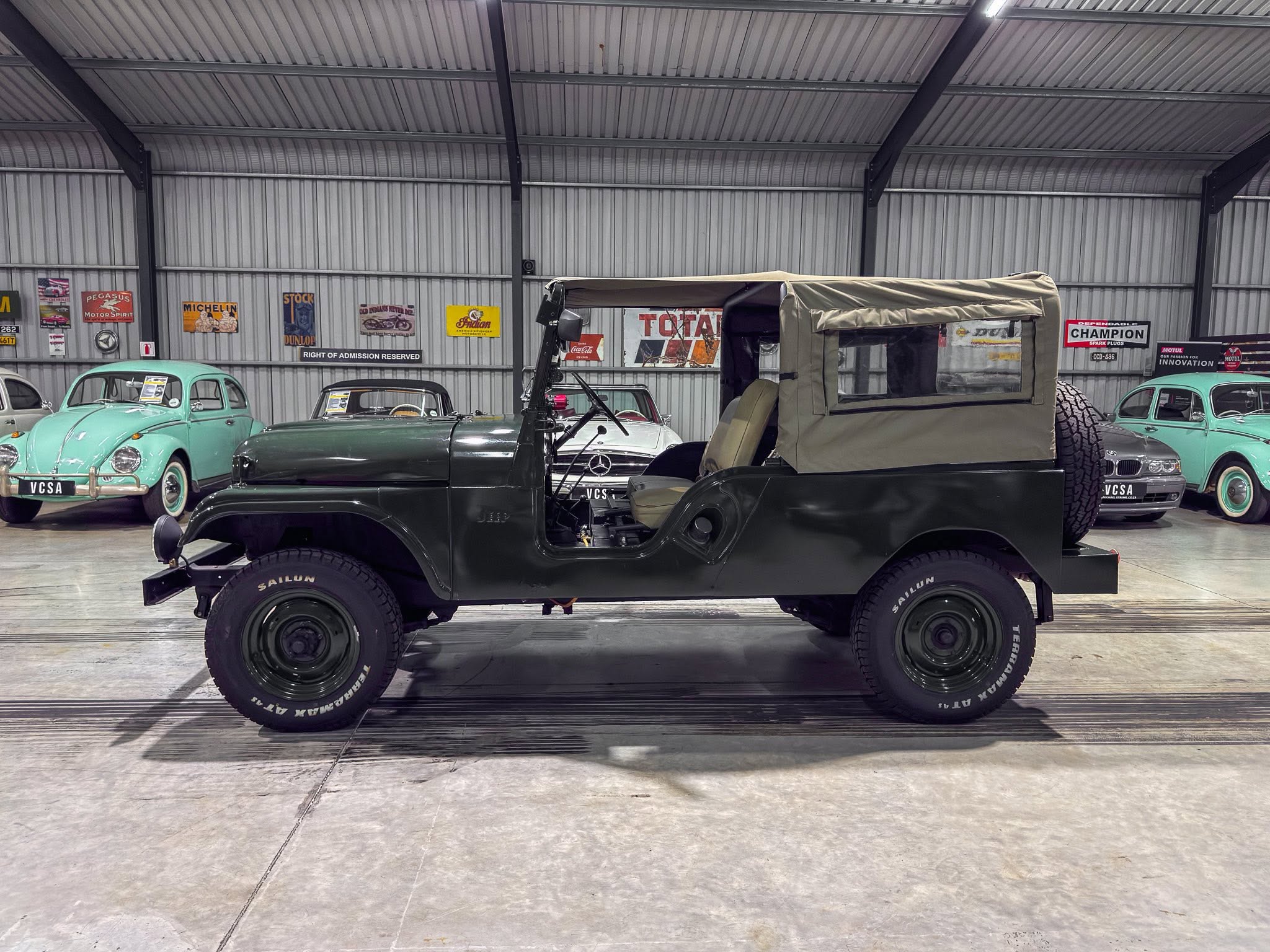Dark green vintage jeep with a beige canvas top parked inside a retro car showroom with classic cars and wall signs in the background