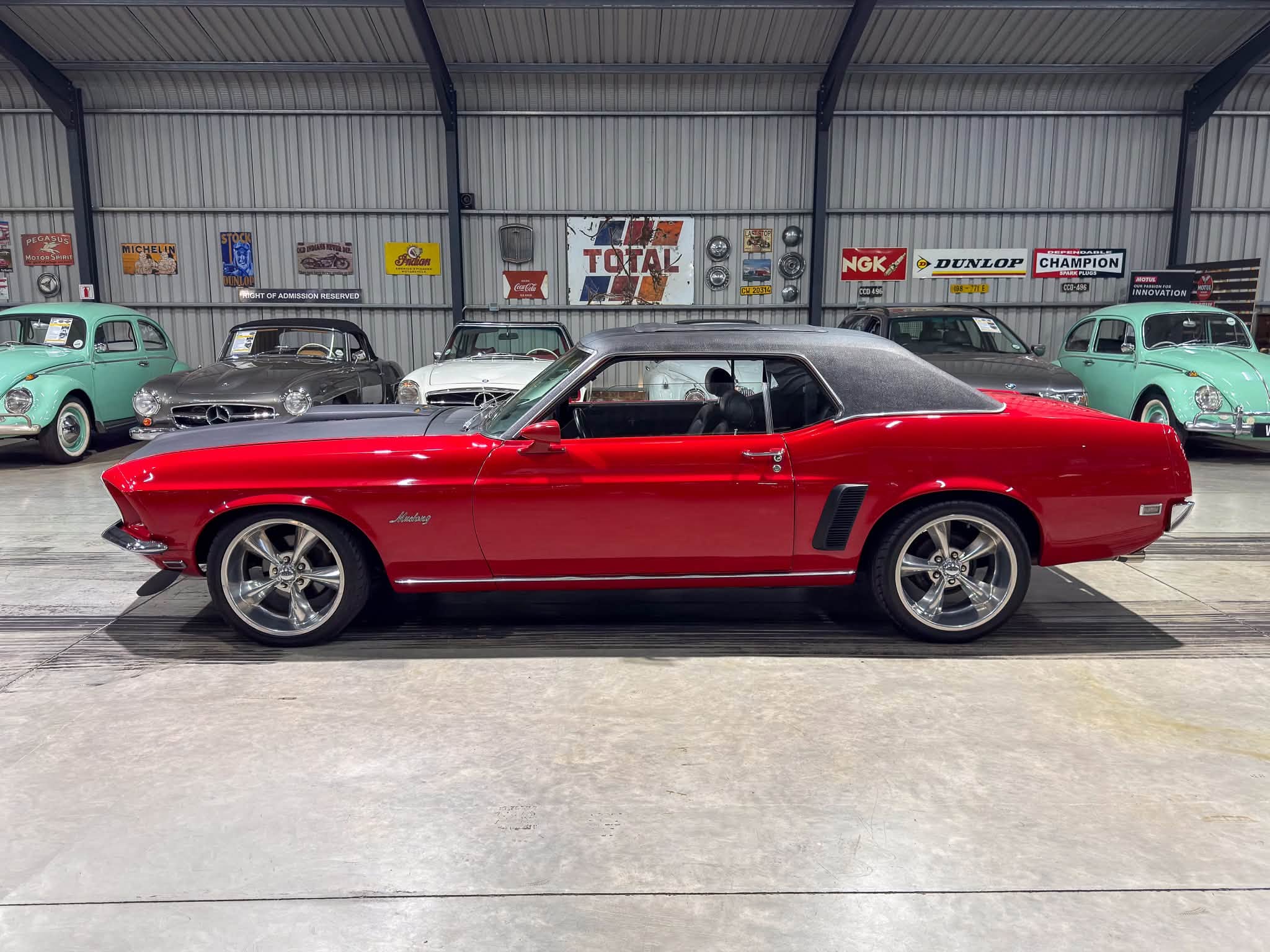 Red classic Ford Mustang with a dark convertible roof, parked indoors among vintage cars and retro signs on a corrugated metal wall.