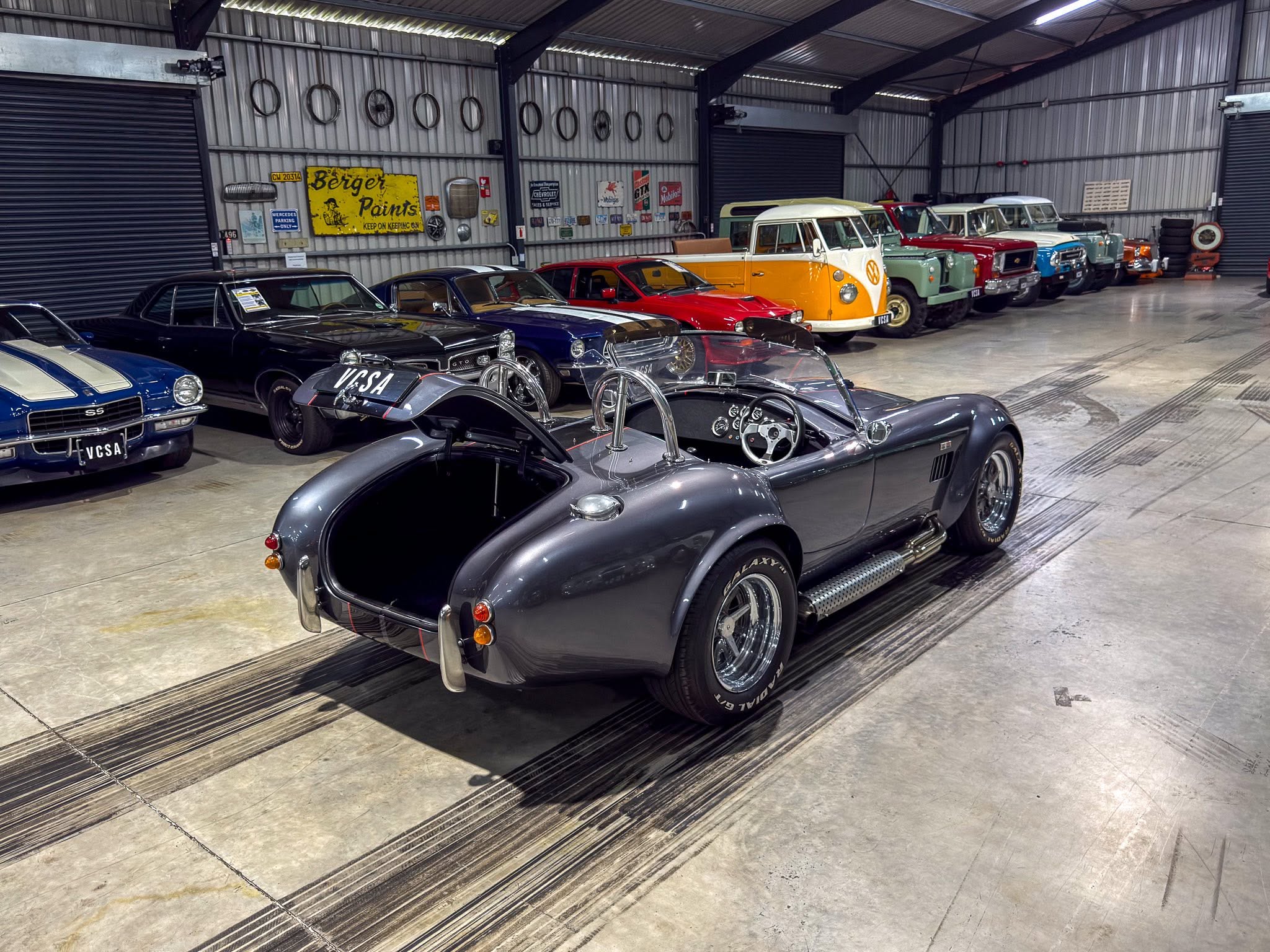 Indoor classic car collection: row of vintage cars in a metal-walled garage with retro signs on the wall, including a yellow and white van in the middle era. Ideally concise and informative for screen readers, background vehicles visible in a showroom-like space.