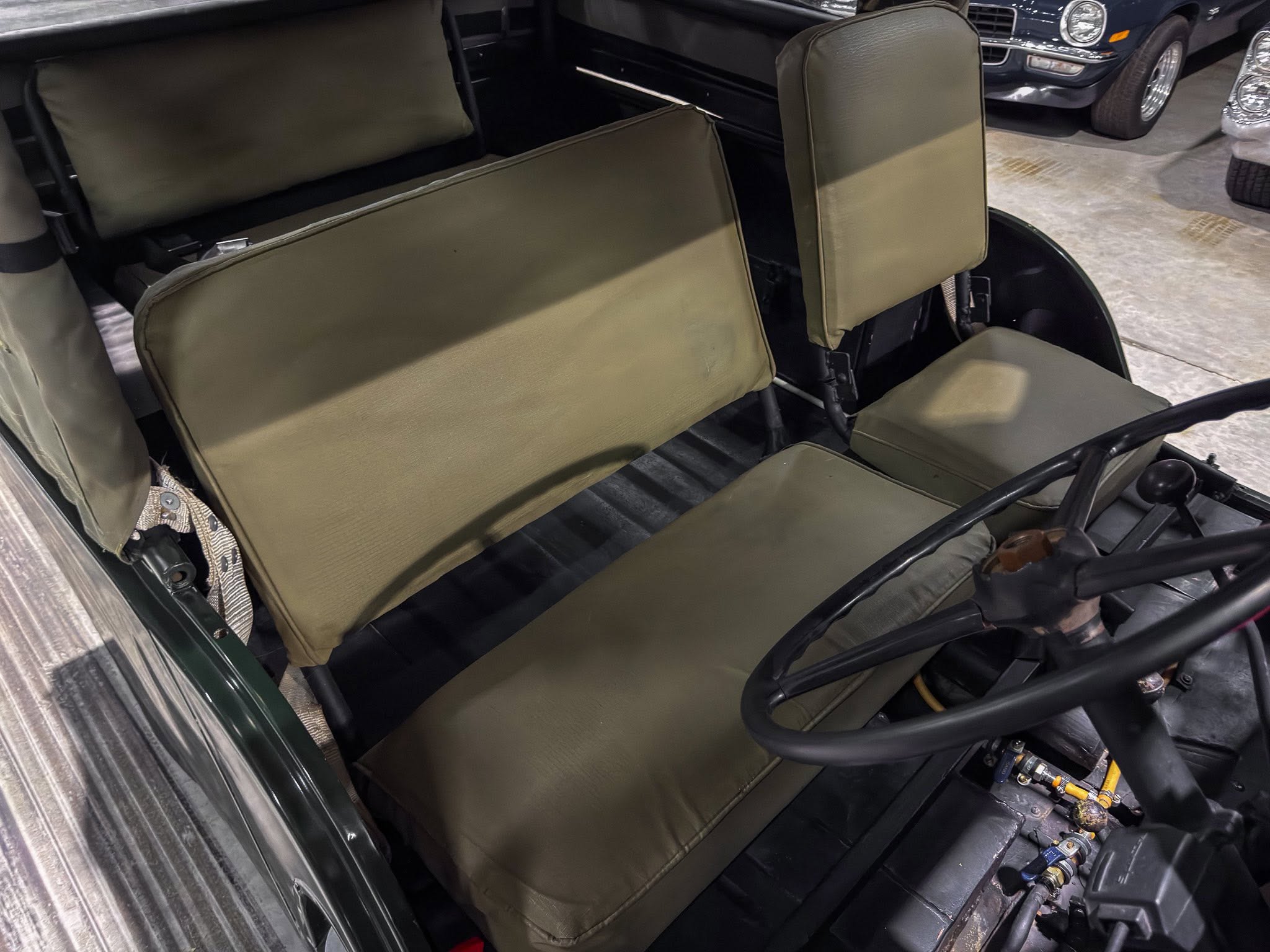 Olive-green vintage car interior with folded front seatbacks and a visible steering wheel on the right side, background car visible outside.