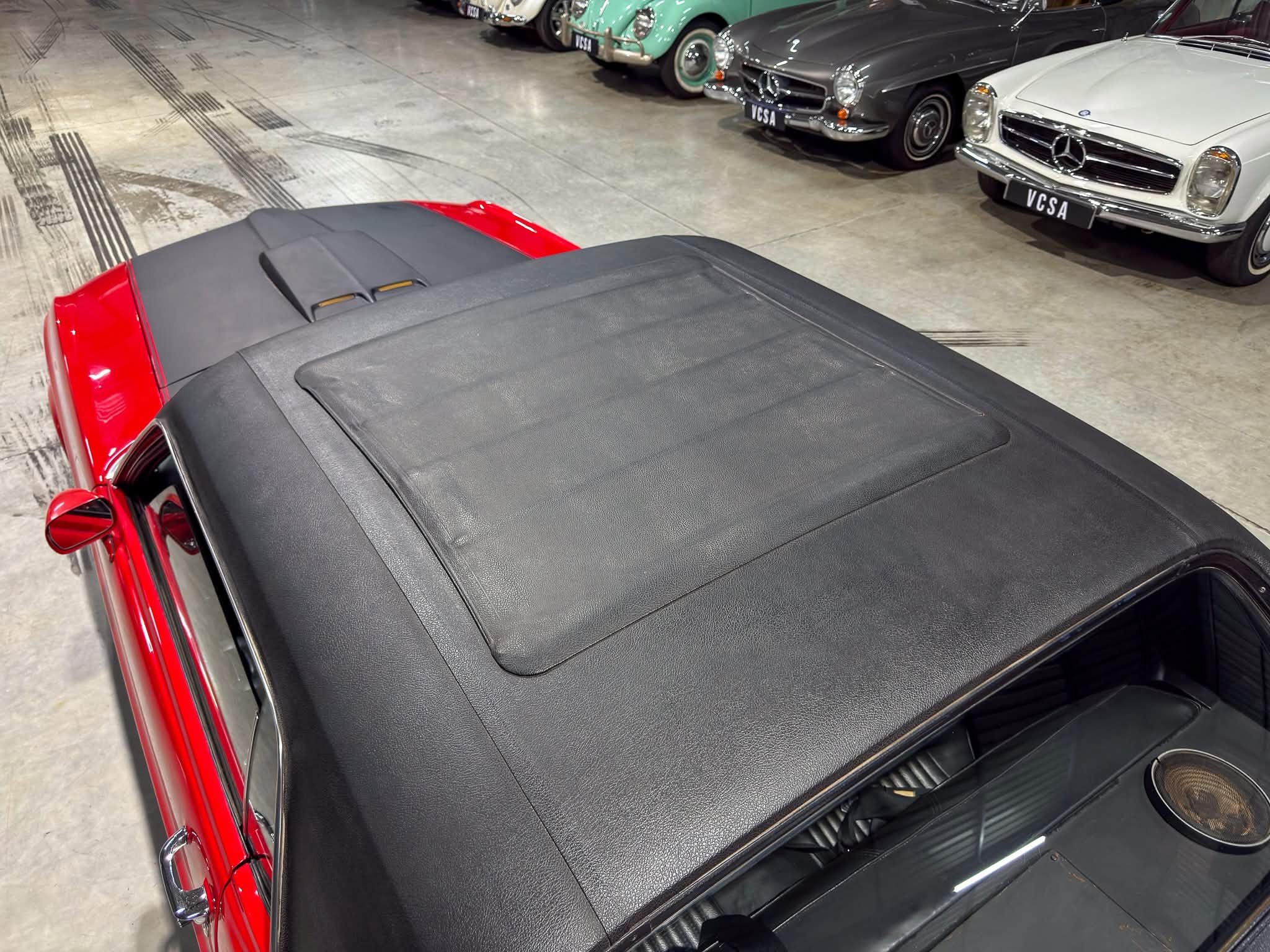 Red vintage convertible with a black soft top, parked in a showroom beside classic cars in the background.