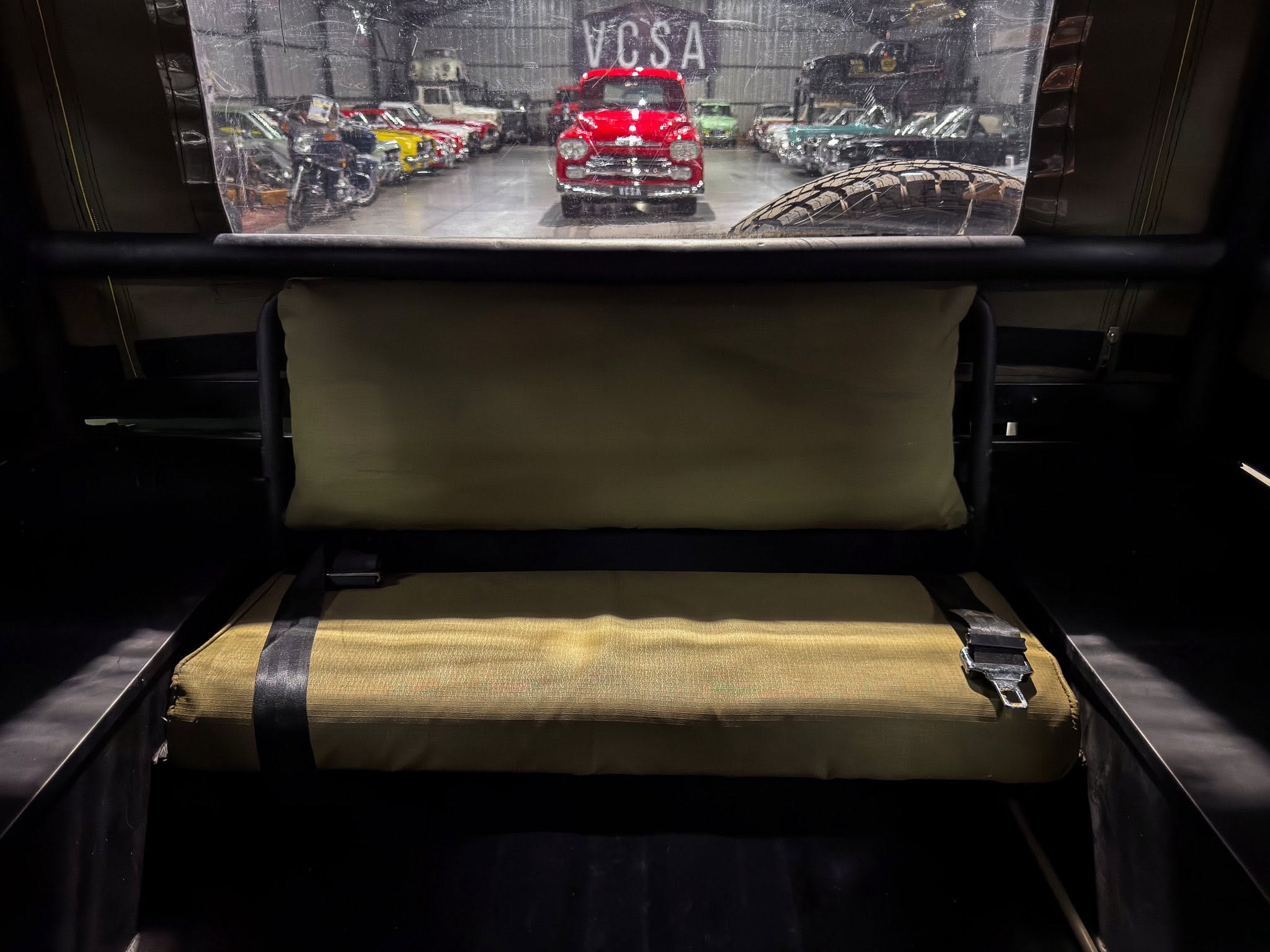 Interior view from the back of a vehicle looking into a classic-car showroom; a red vintage car is centered with other cars along the sides and a large tire visible on the right face of the scene.