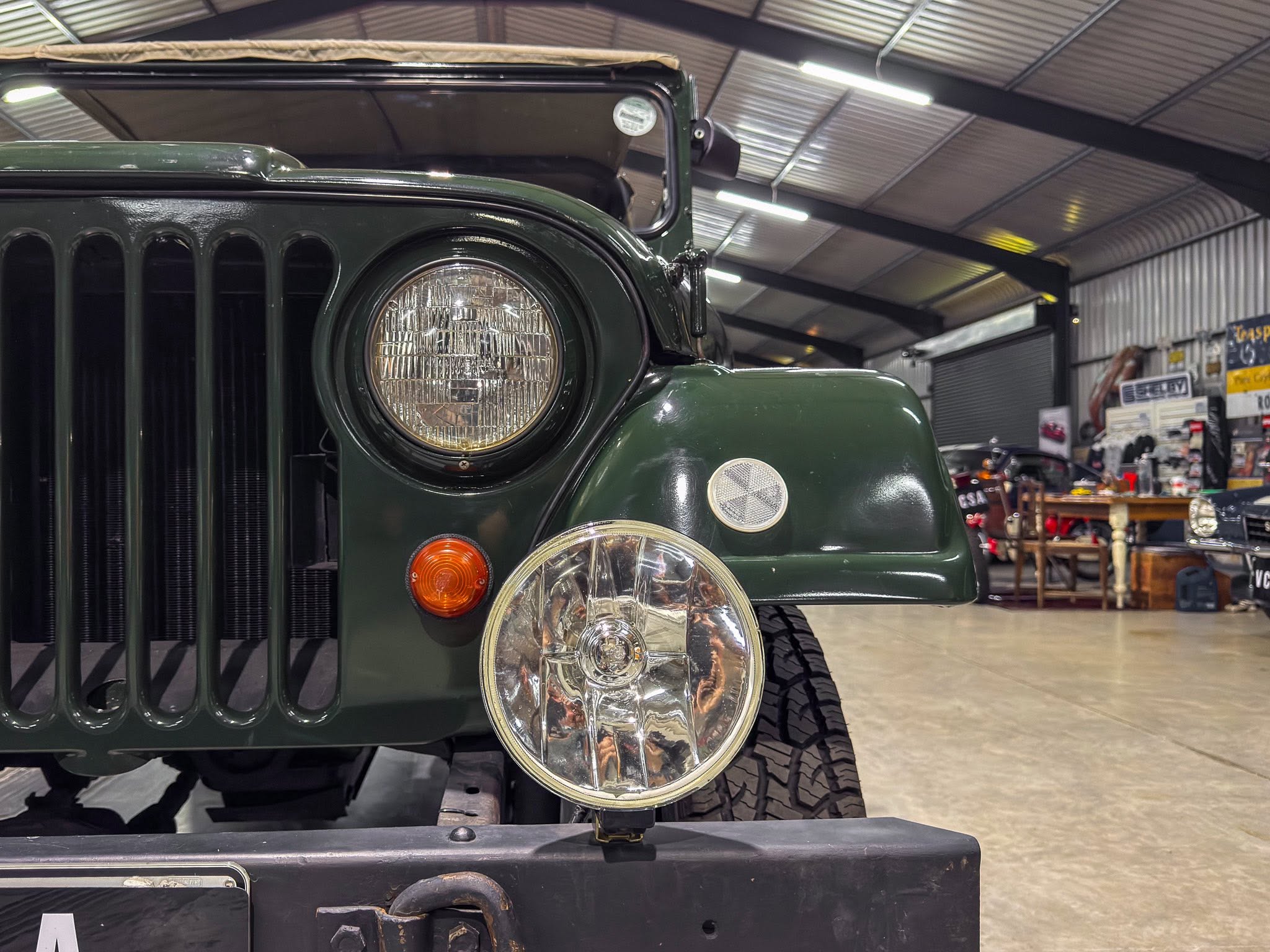 Close-up of the front left corner of a dark green vintage Jeep, highlighting a round headlight, vertical grille slots, and robust bumper in a showroom.