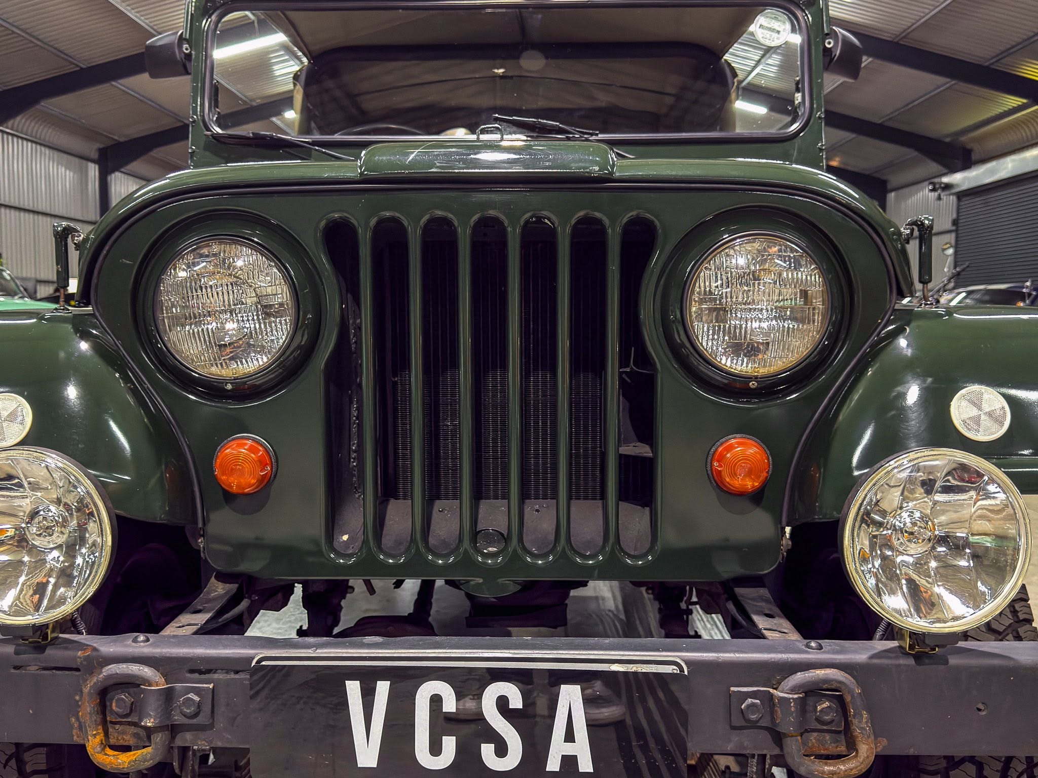 Front view of a dark green vintage Jeep with round headlights, vertical grille slots, and a license plate reading VCSA.