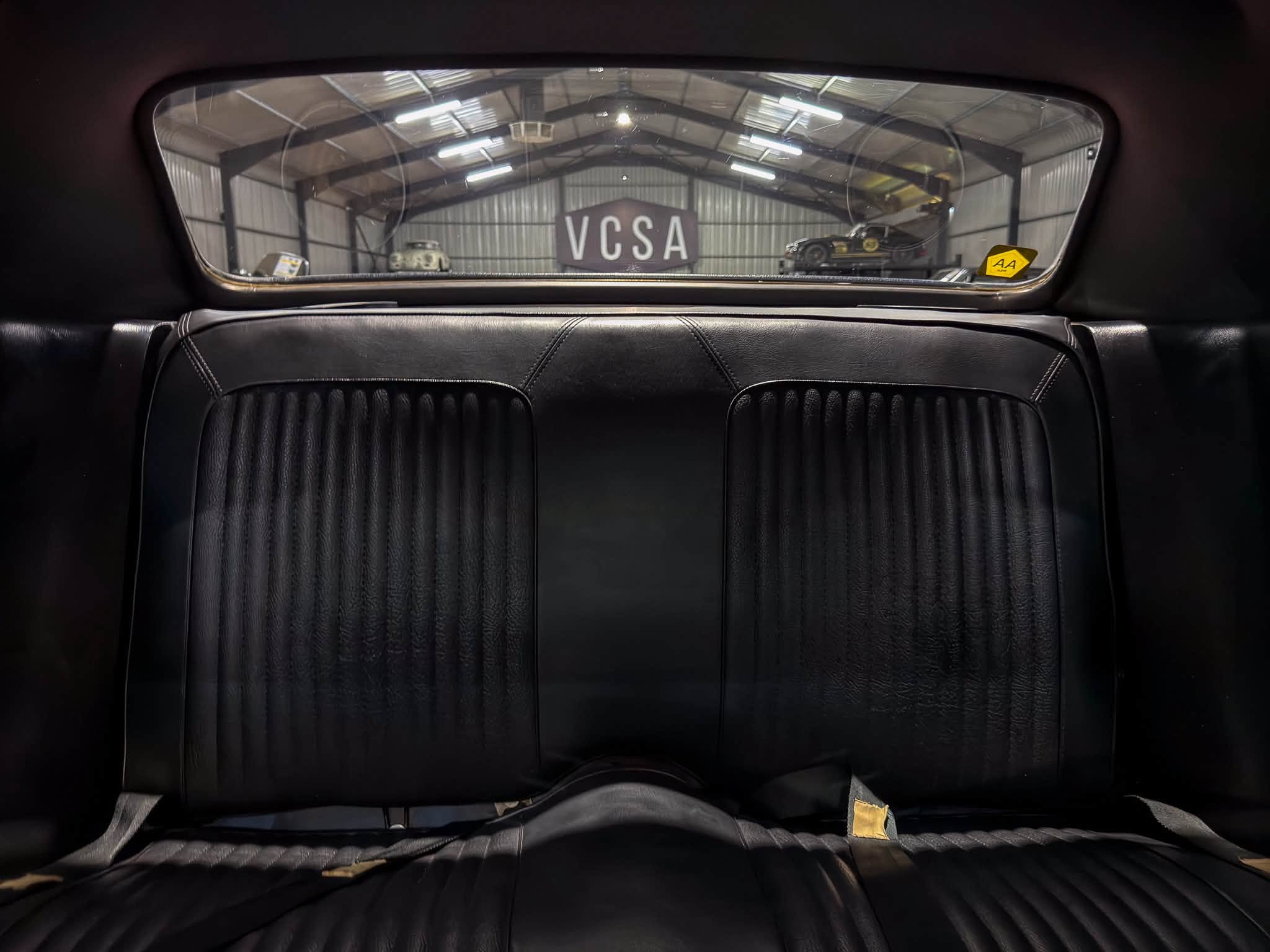 View from the backseat looking through a car's rear window at a warehouse with a large 'VCSA' sign and vintage cars inside. Leather seats frame the image.