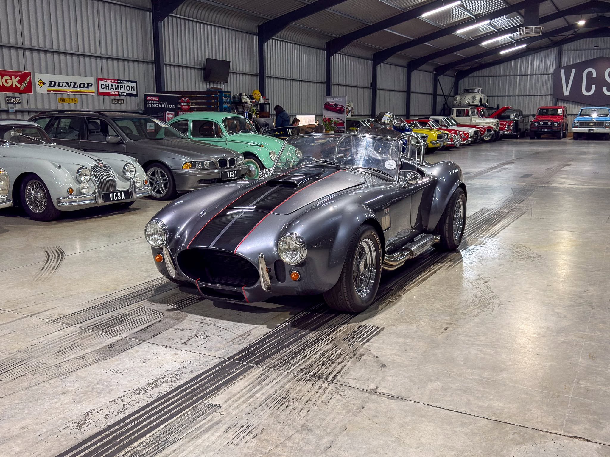 Indoor showroom filled with vintage and classic cars parked in a row, foreground features a gray sports car with red stripes.