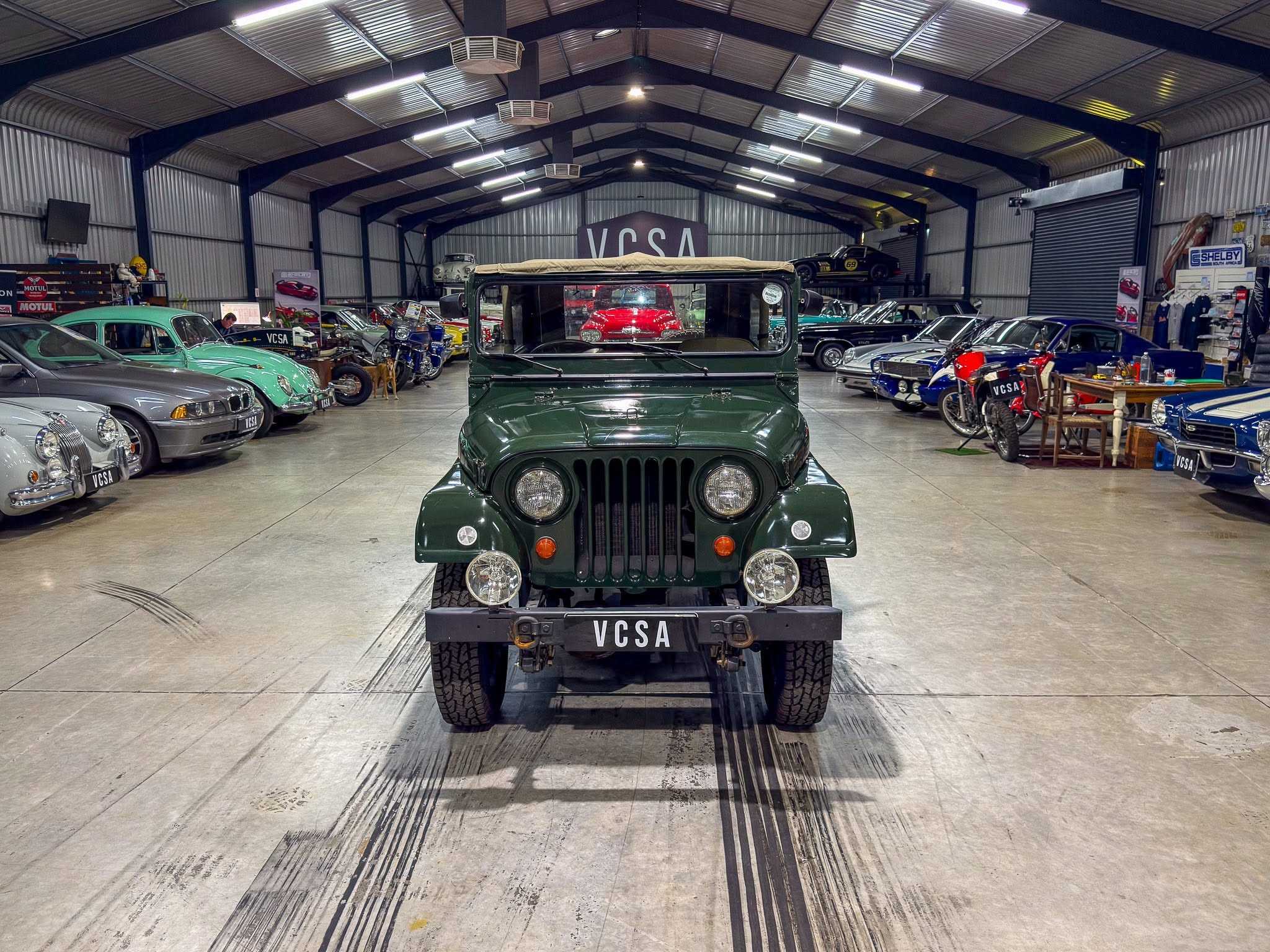 Indoor classic car showroom with a green vintage jeep centered in the foreground and rows of antique cars along the sides, VCSA license plates visible.