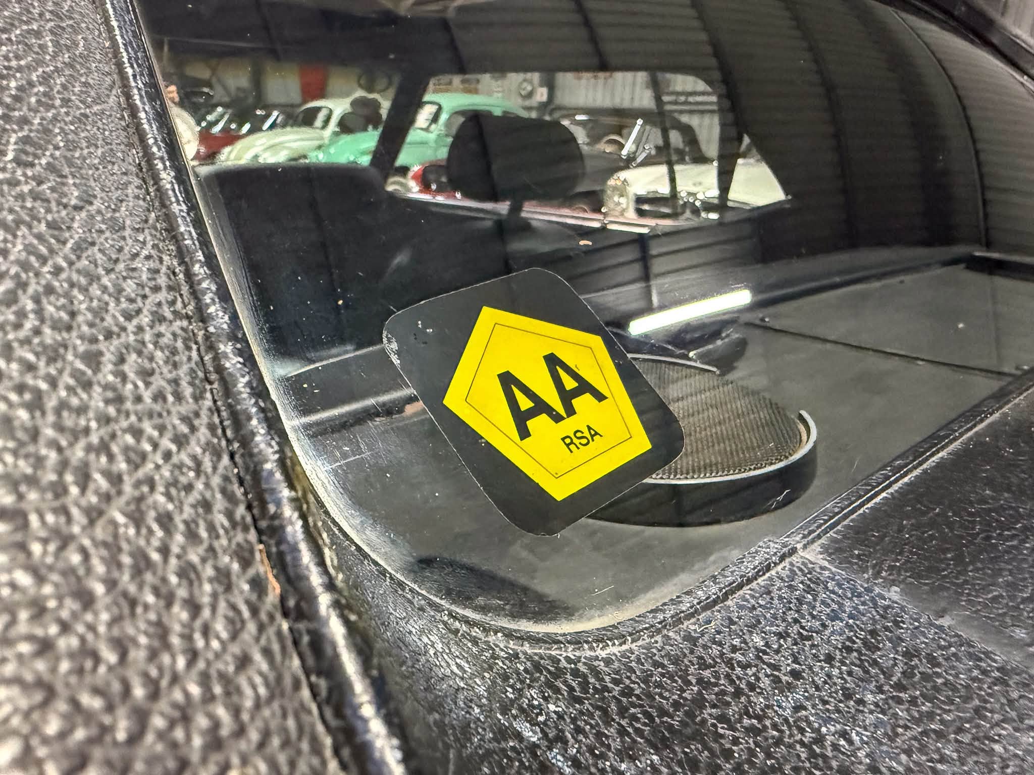 View through a car windshield showing a yellow caution sign on the dashboard and a workshop with vintage cars in the background.
