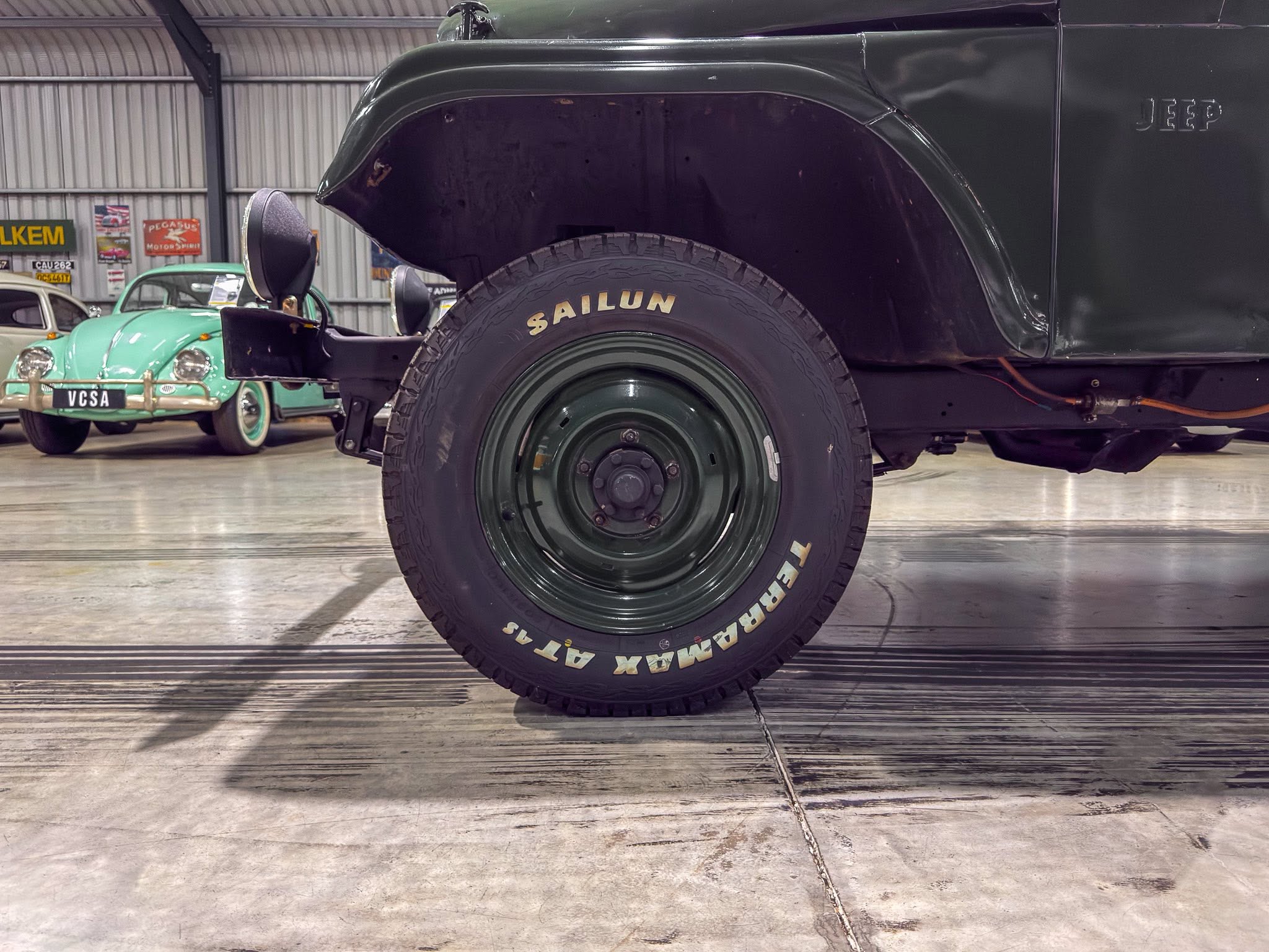 Close-up of a large dark-green vintage truck’s front wheel and fender, with a mint-green classic car visible in the background inside a museum.