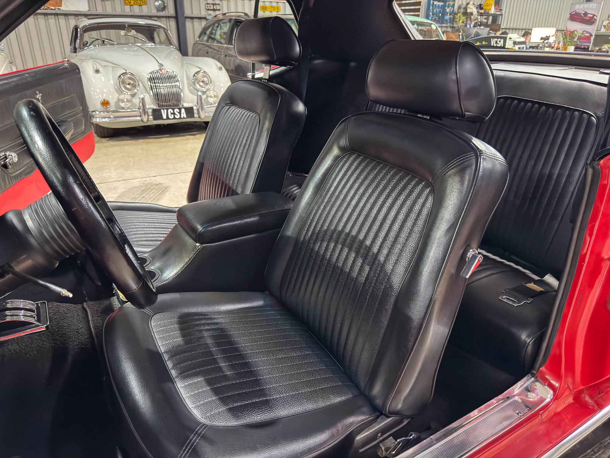 Red vintage car interior with black leather seats and red door frame; showroom with classic cars visible outside edge of image.