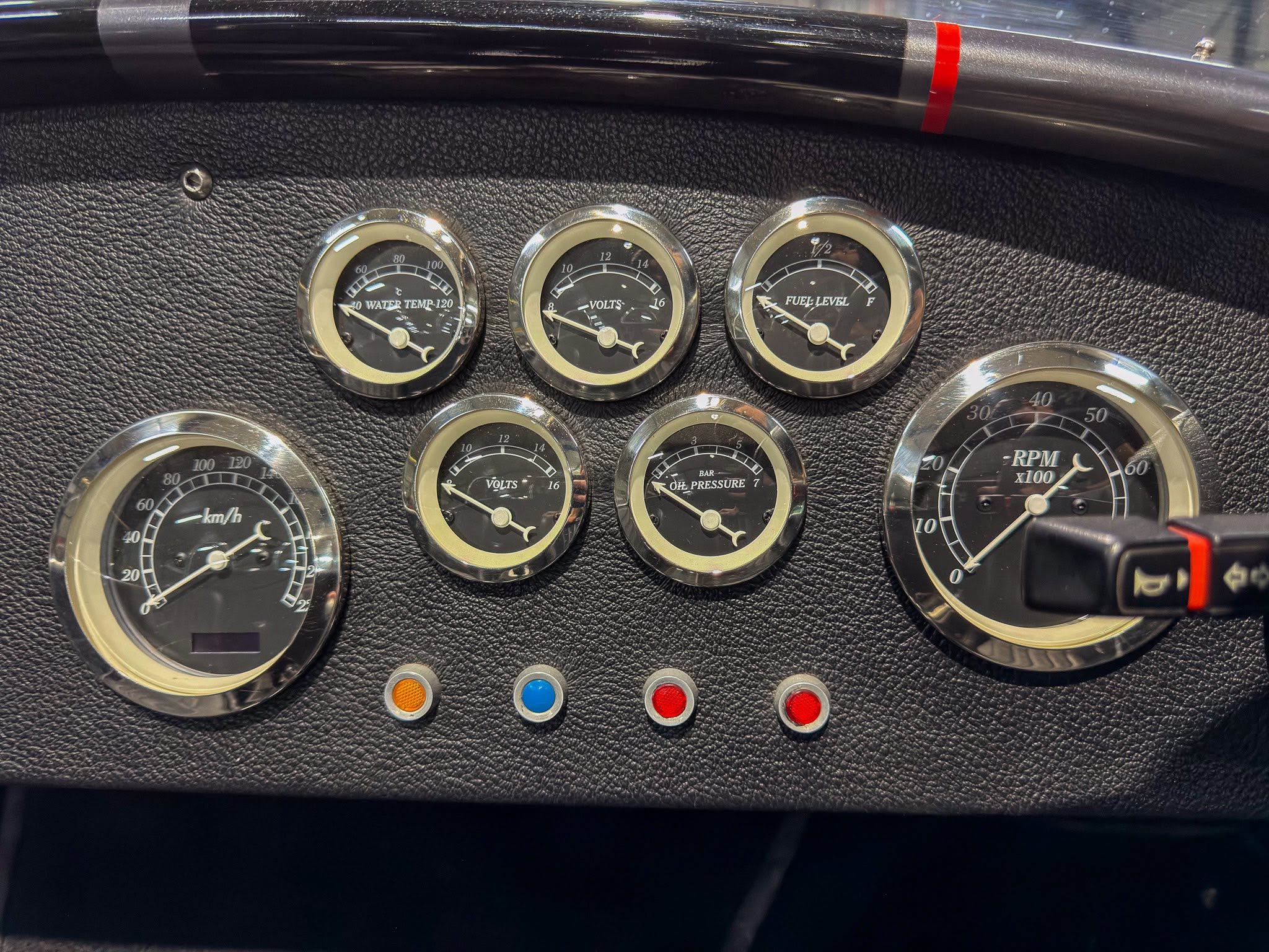 Close-up of a vintage car dashboard with chrome-rimmed gauges for water temperature, volts, and fuel level on a black textured panel.