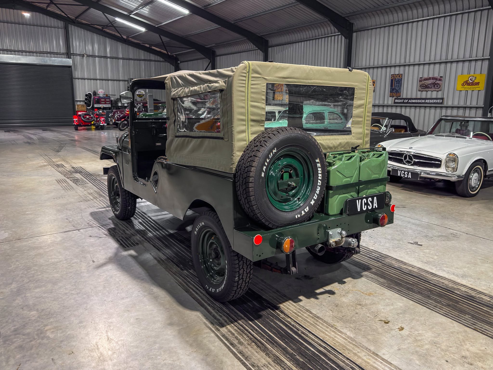 Green vintage military jeep with tan canvas cover and spare tire mounted on the back, parked inside a metal-walled showroom with classic cars and retro signs in the background.