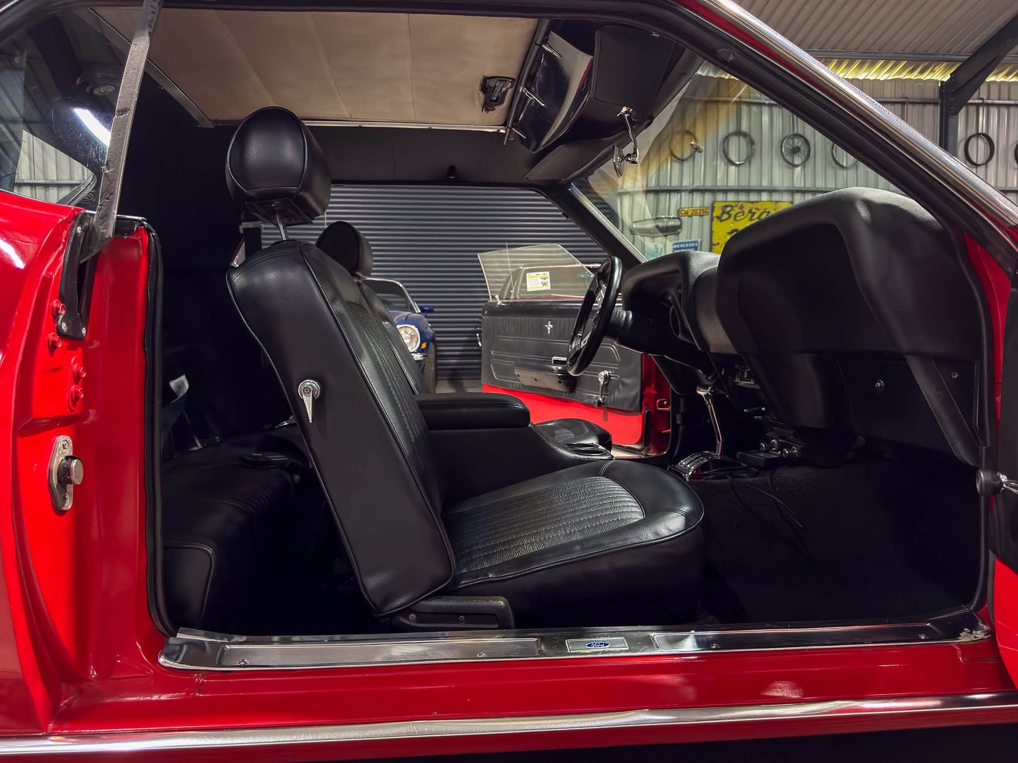 Open red car door reveals black leather front seats and a glimpse of the dashboard inside a garage or workshop edge visible in background.