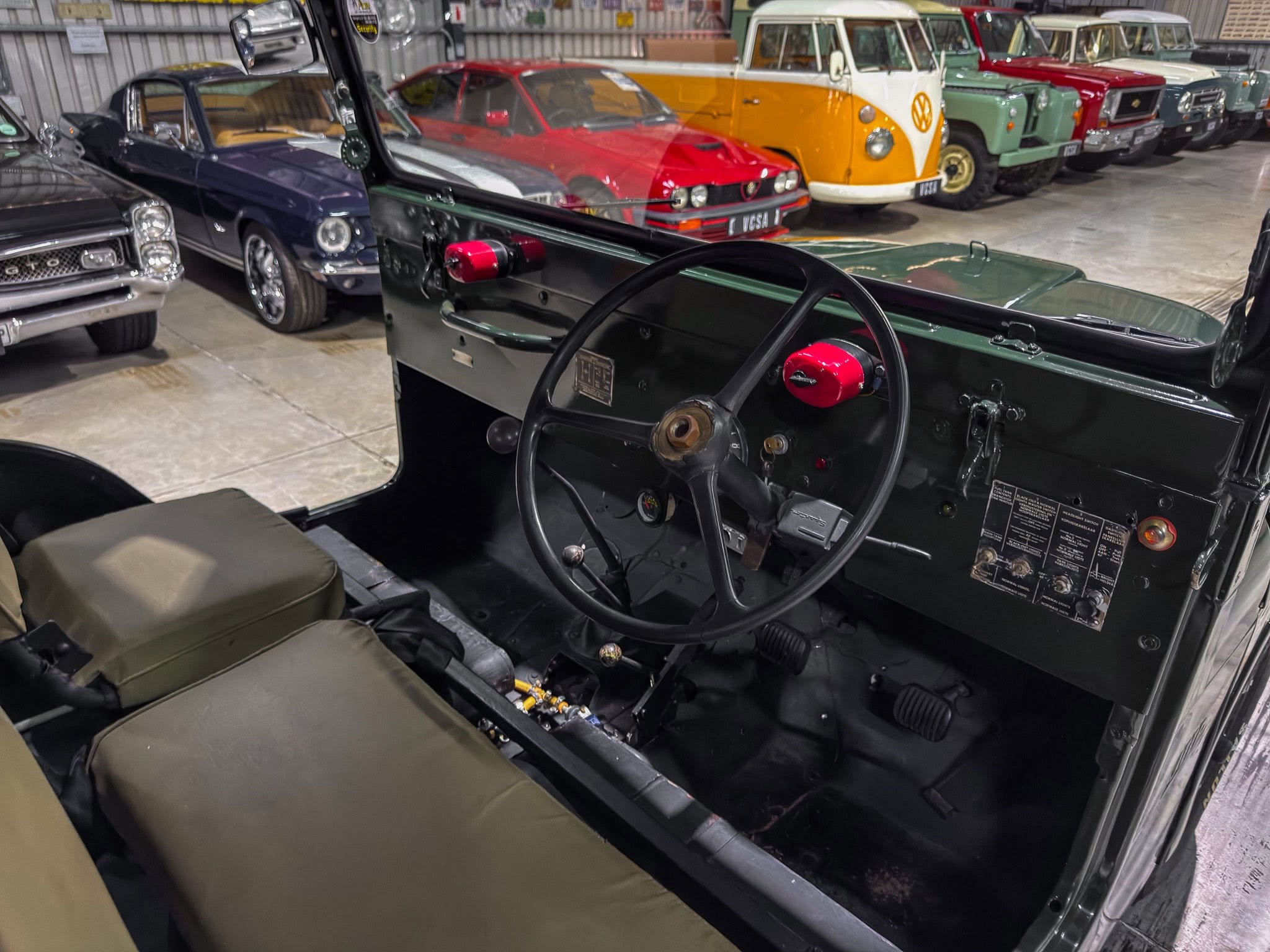 Interior view of a vintage open-top car: black dashboard, large steering wheel, red knobs, with classic cars visible in a showroom background.