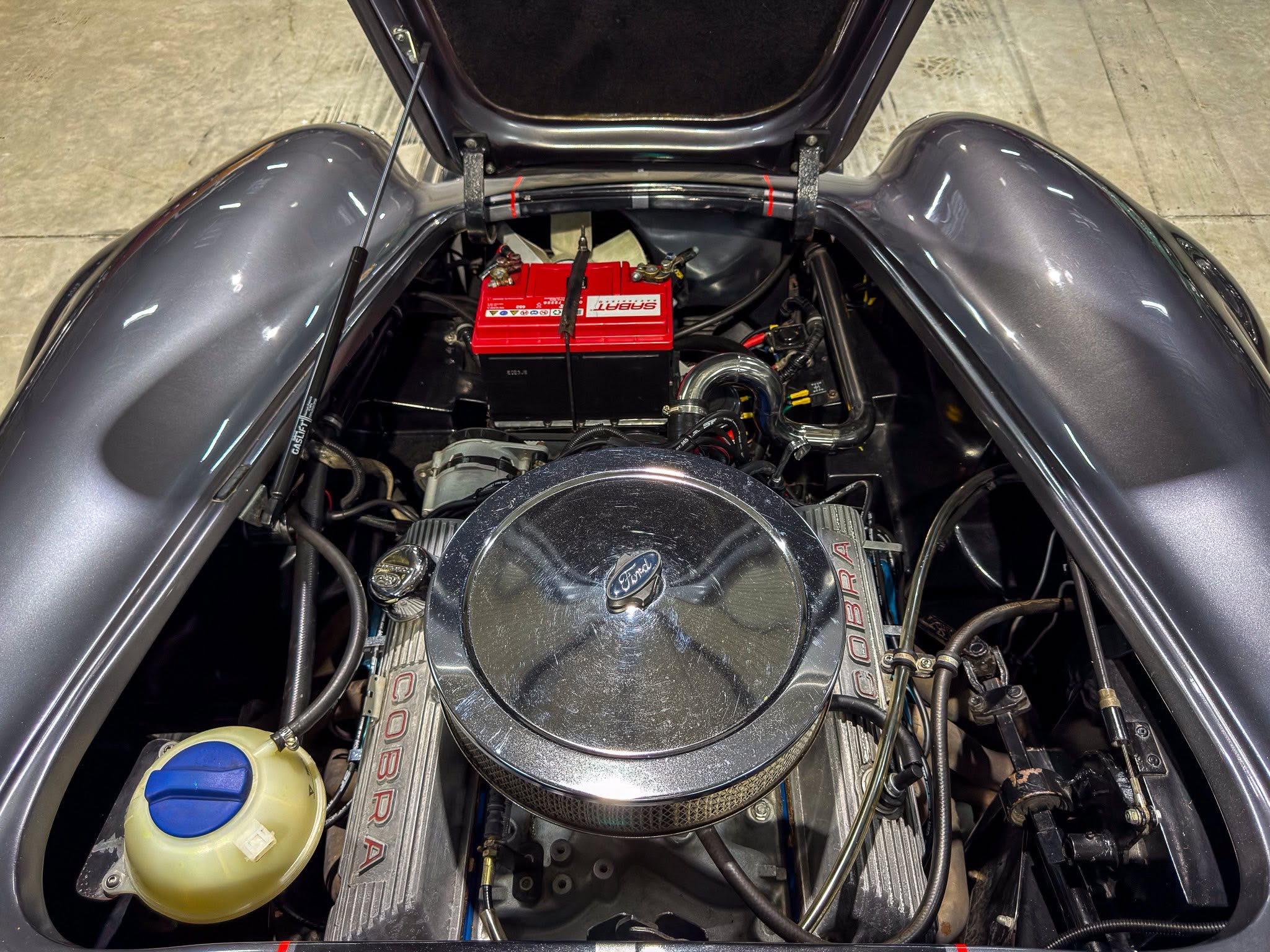 Top-down view of a car engine bay with a red battery, chrome air cleaner, and various hoses and wires visible beneath the hood light.