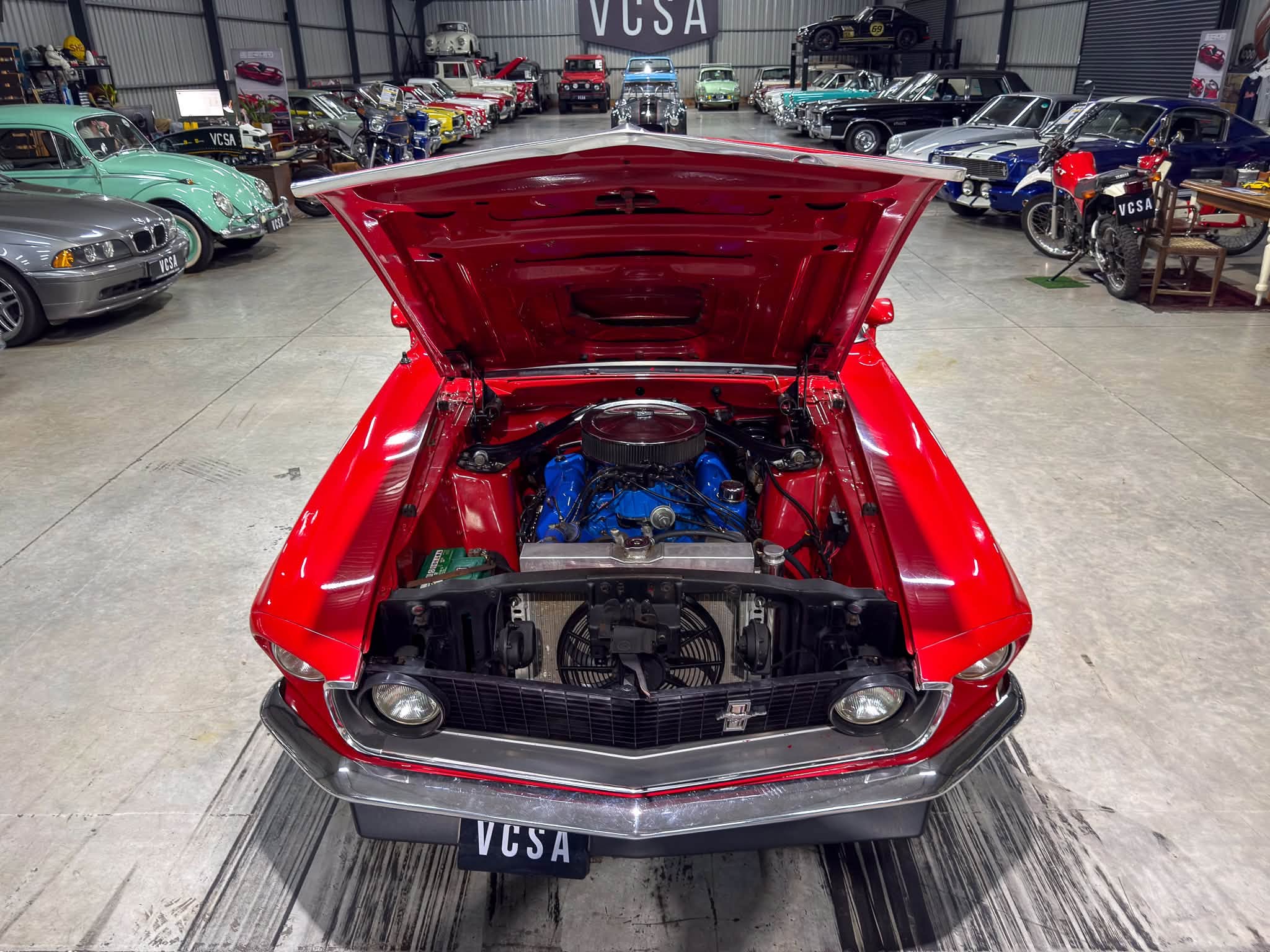 Red vintage car with its hood open, blue engine exposed, in a showroom lined with classic cars in the background.