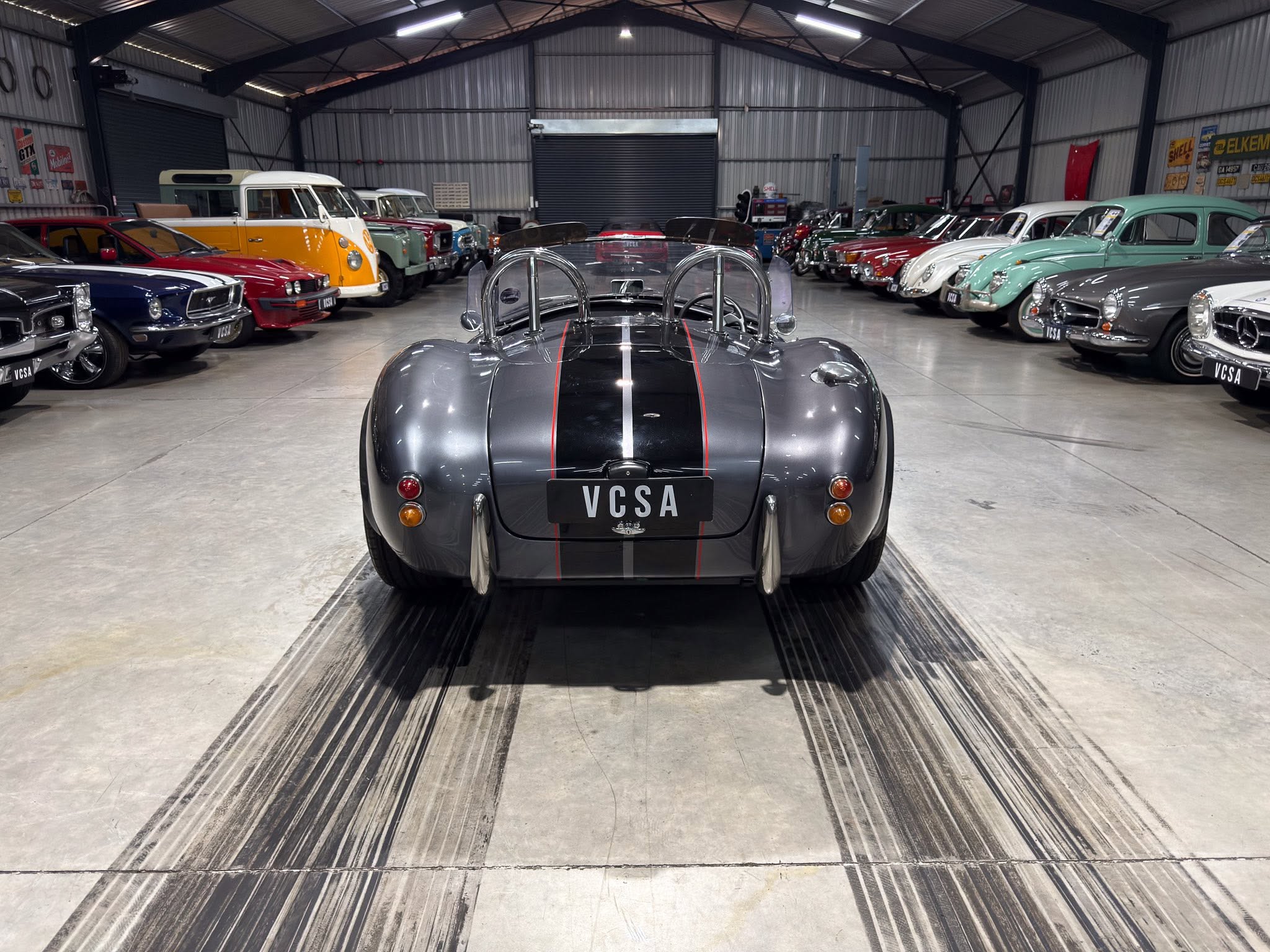 Indoor showroom or warehouse filled with vintage cars lined up along both sides; a gray vintage roadster with red racing stripes is centered in the foreground, viewed from behind.