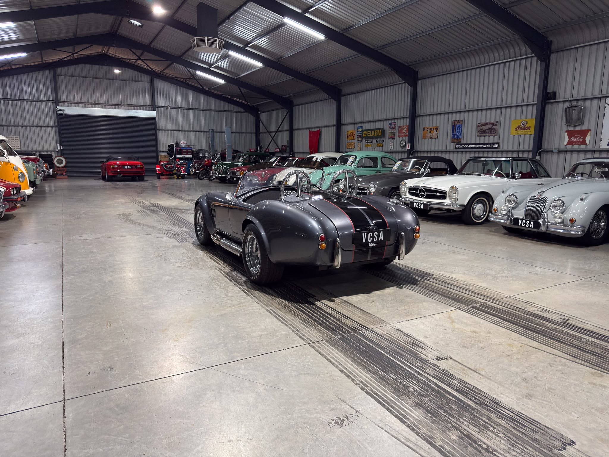 Indoor classic car showroom with a row of vintage cars, including a gray open-top racer in the foreground.