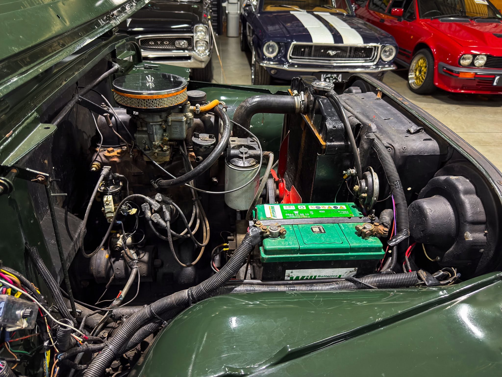 Open engine bay of a vintage green car with exposed hoses, air filter, and wiring; classic cars visible in the background.