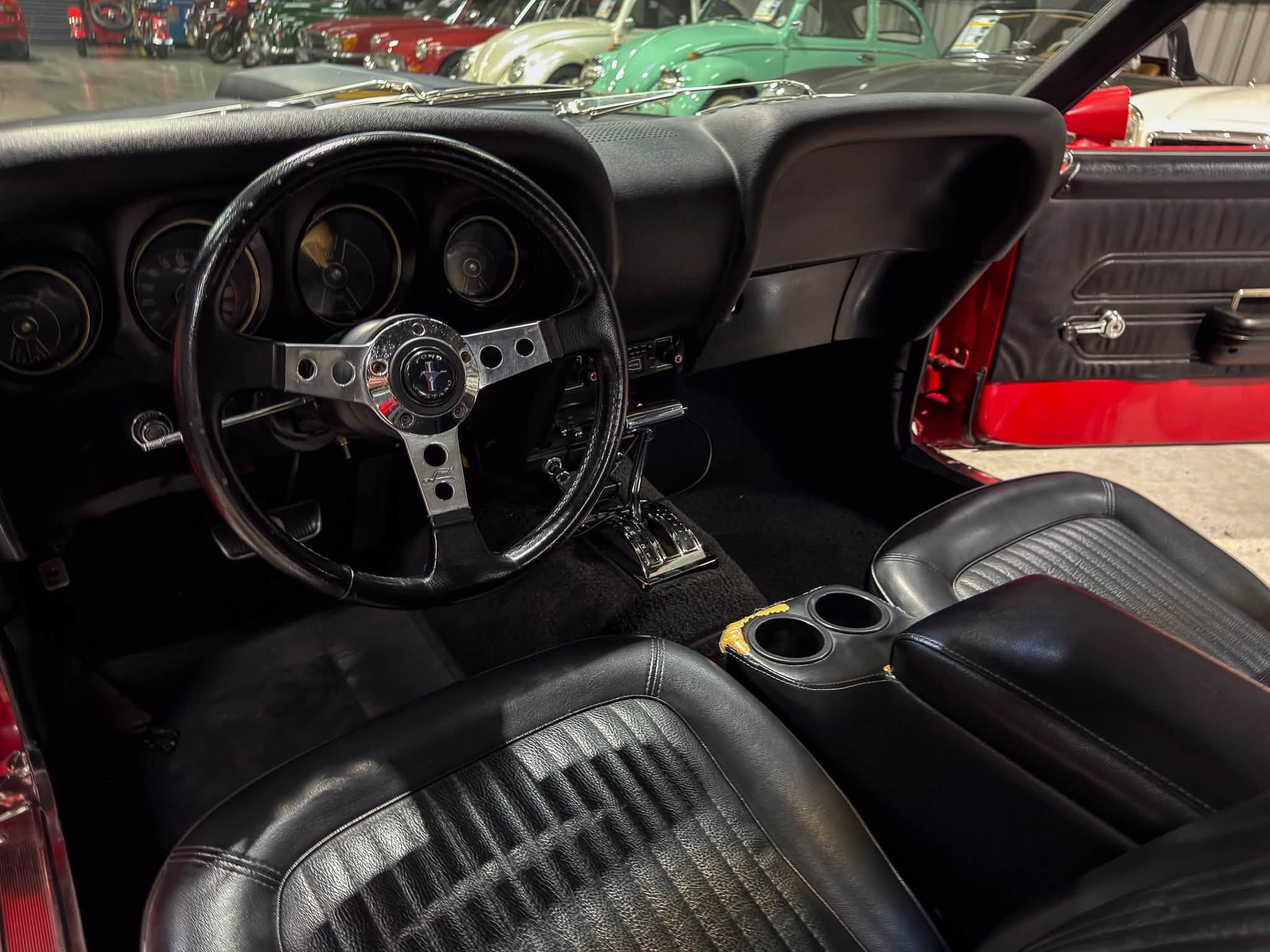 Interior view of a black vintage Mustang: chrome steering wheel, analog gauges, and a red door panel in a classic car showroom