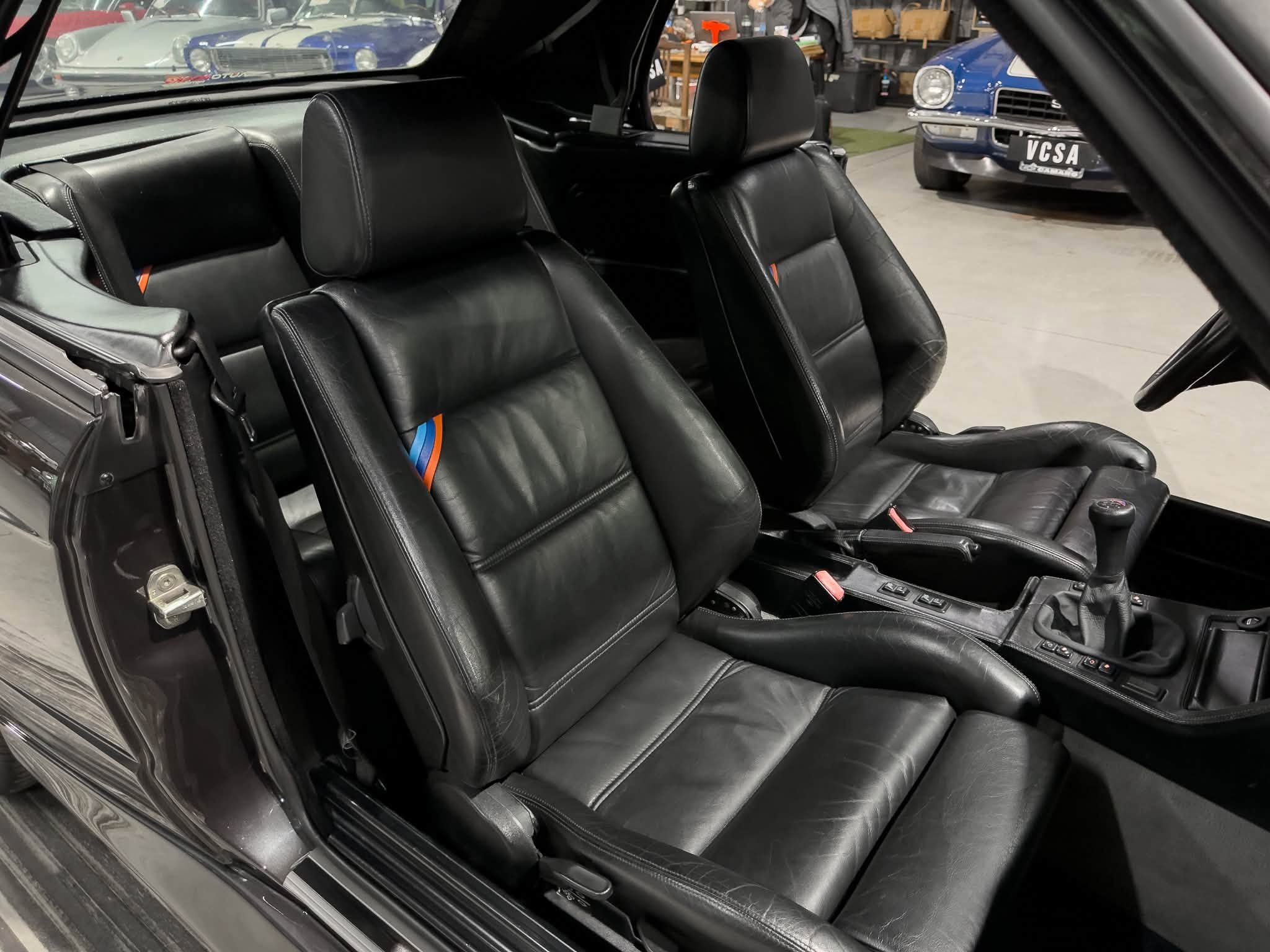 Inside a black leather car cabin with front seats and center console, showroom of classic cars visible through the windows outside.