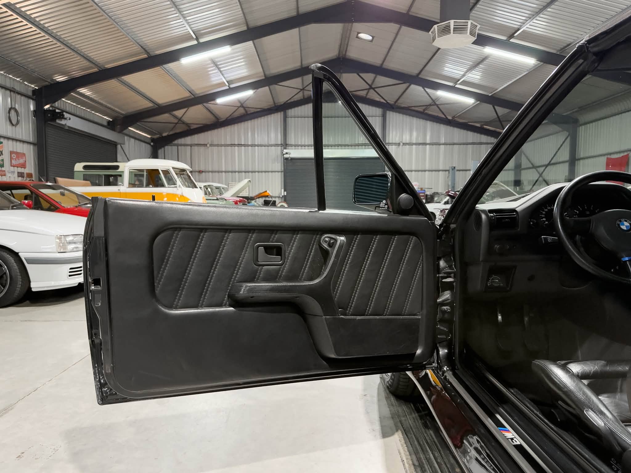 Open driver's-side door of a black BMW, exposing the black leather interior and steering wheel, with a warehouse full of vintage cars in the background.