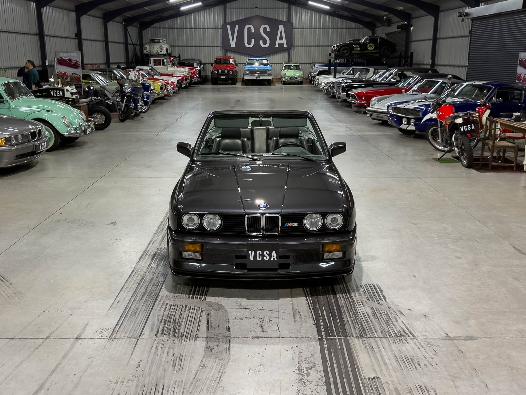 Front view of a black BMW M3 convertible in a spacious classic car showroom, with rows of vintage cars on both sides and a VCSA sign above.