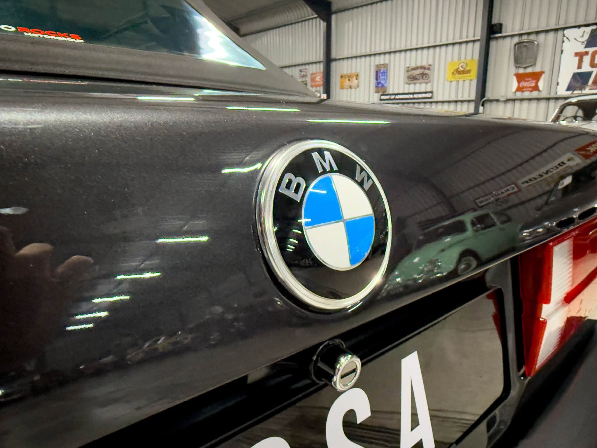 Close-up of the BMW badge on a black car hood inside a workshop with posters on the corrugated metal walls in the background.