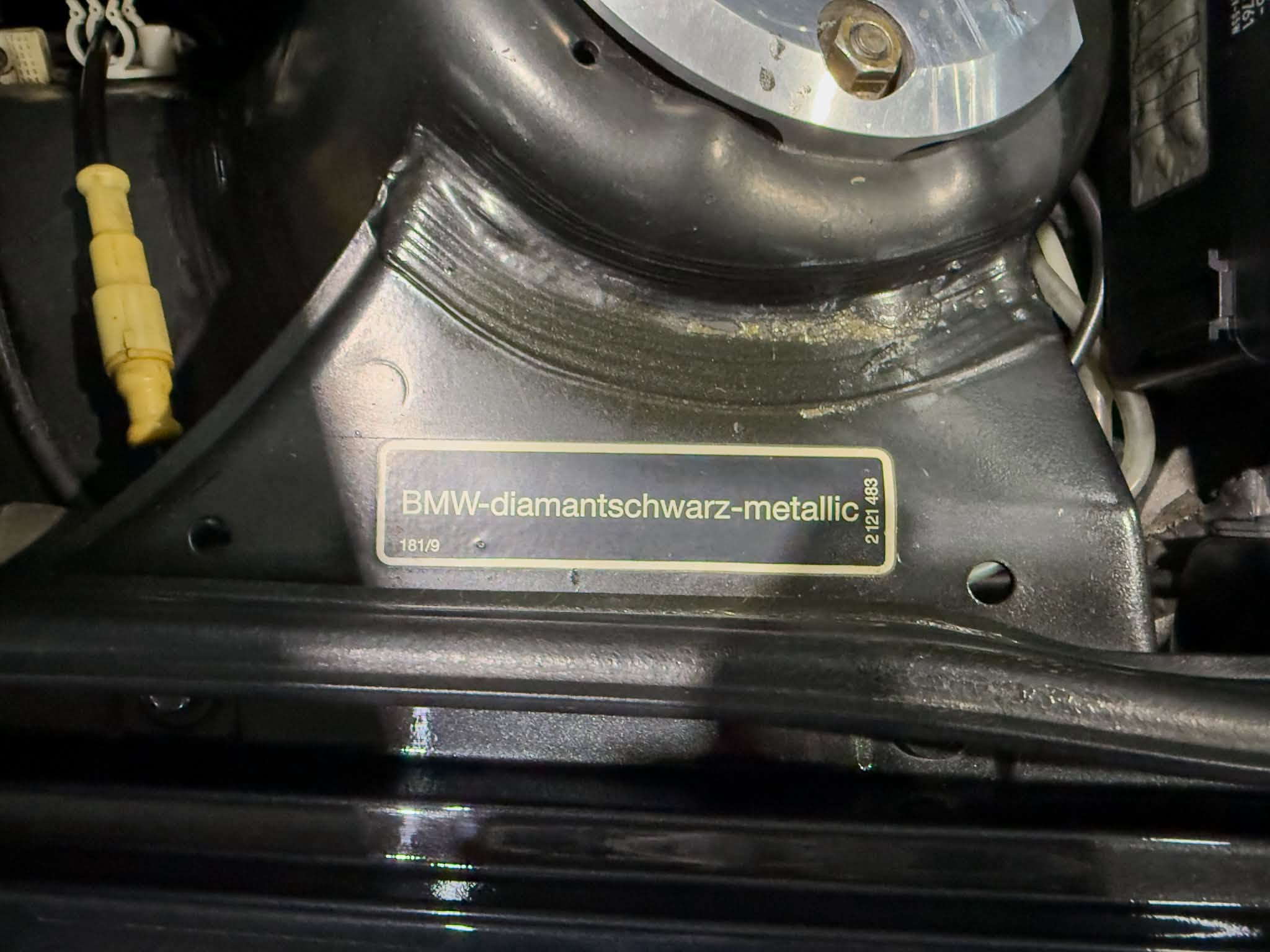 Close-up of a vehicle engine component with a gold-bordered plate reading 'BMW-diamantschwarz-metallic' and surrounding black metal parts.
