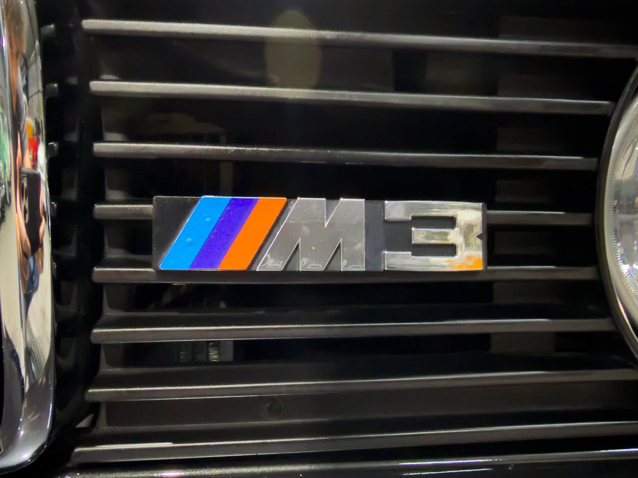 Close-up of a black car grille with a blue, purple, and orange M-style stripe badge prominent in the lower left area.
