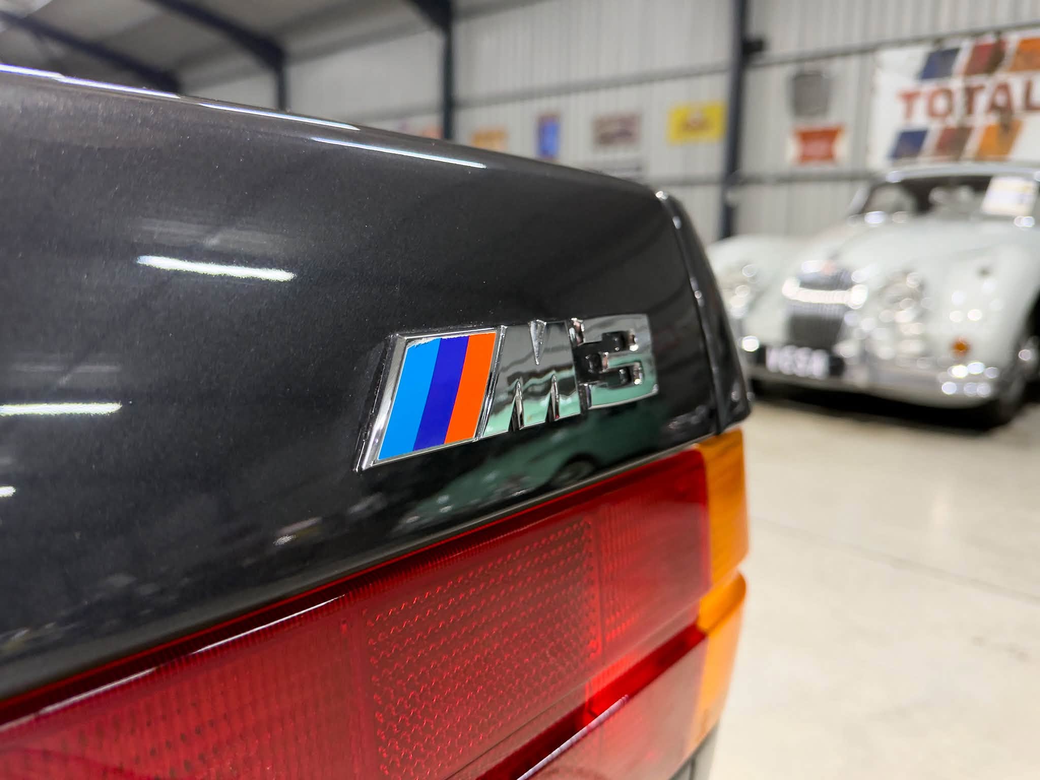 Close-up of a dark car rear with the BMW M badge and red tail light in a showroom.`Note: remove backtick in actual output`