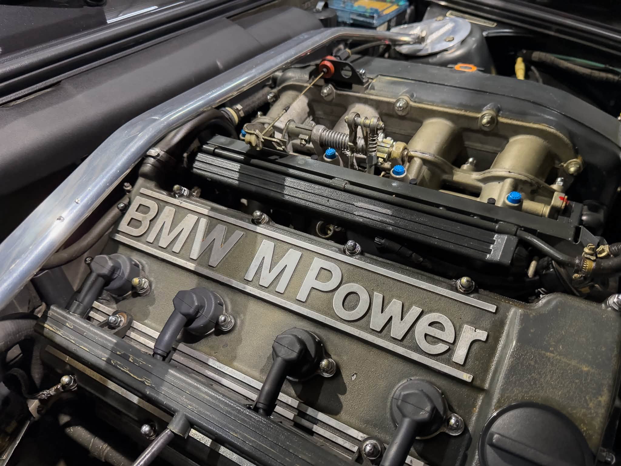 Close-up of a BMW engine bay with the 'BMW M Power' valve cover and mechanical throttle linkage.
