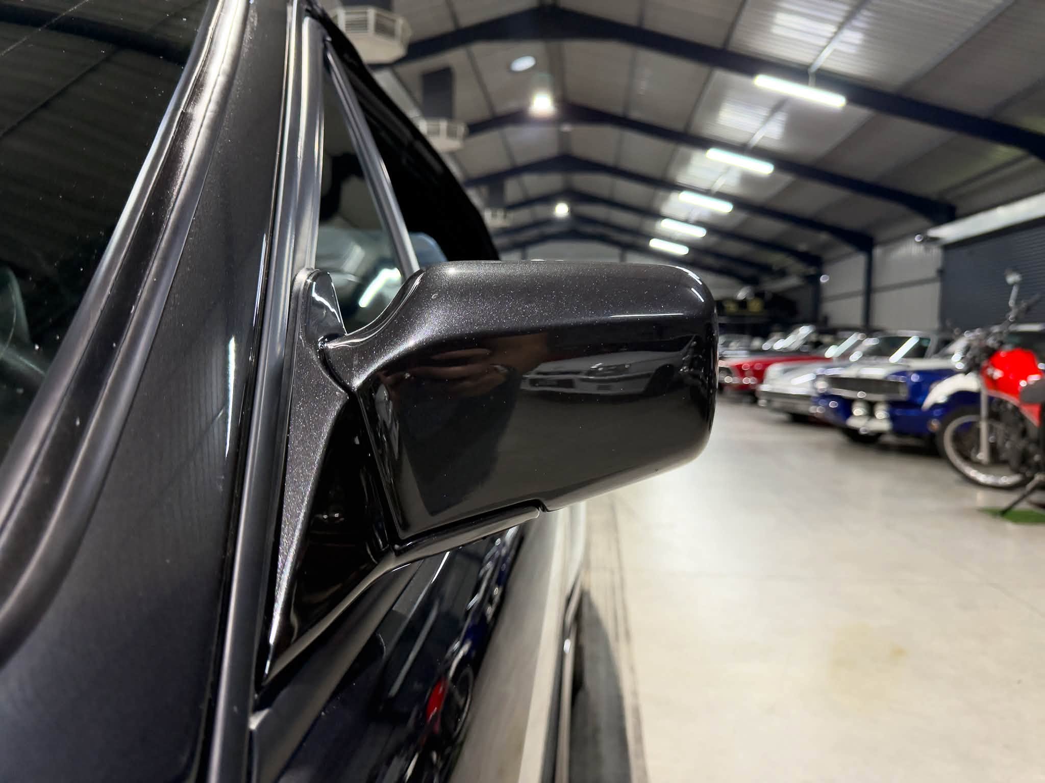 Close-up of a glossy black car side mirror in a showroom with other cars in the background.