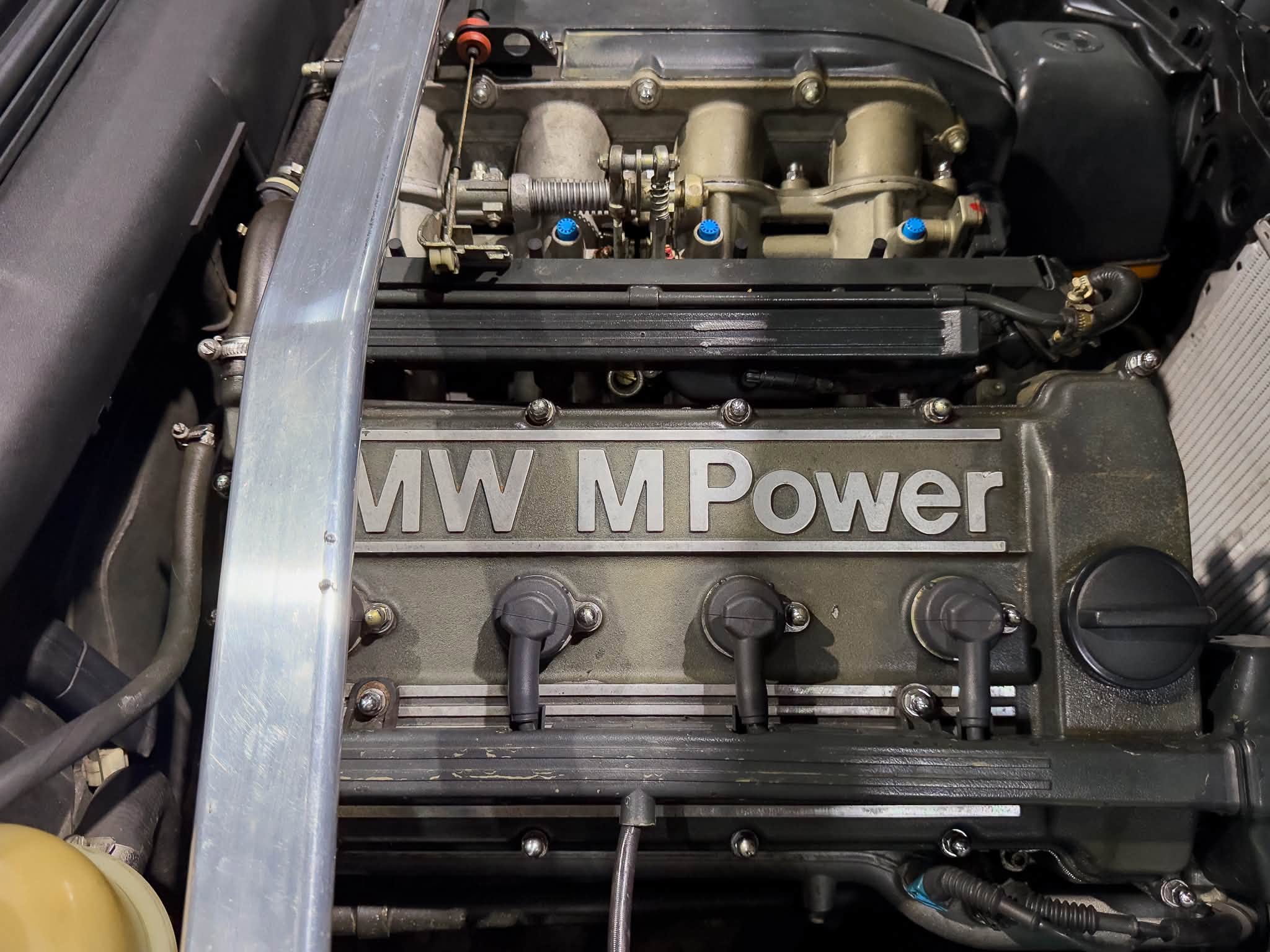 BMW engine bay with 'M Power' badge, blue valve caps, and exposed engine components
