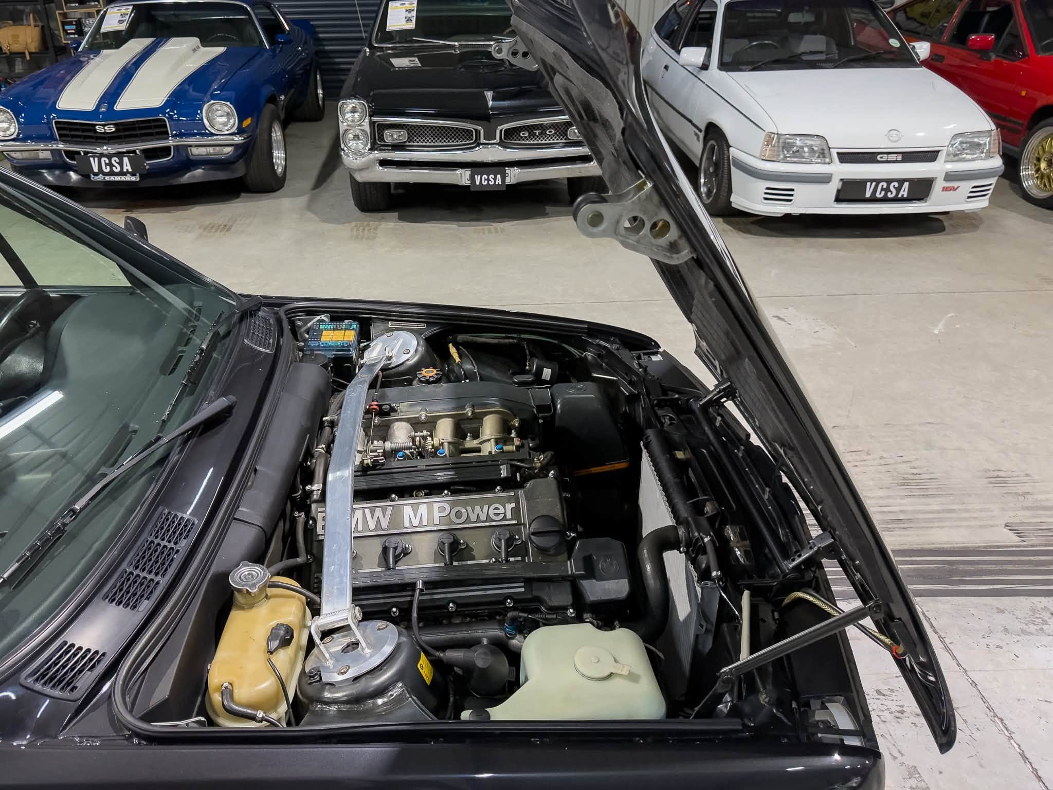 View from inside a car with the hood open, showing a BMW engine labeled 'BMW M Power' in a garage with blue and white classic cars in the background.