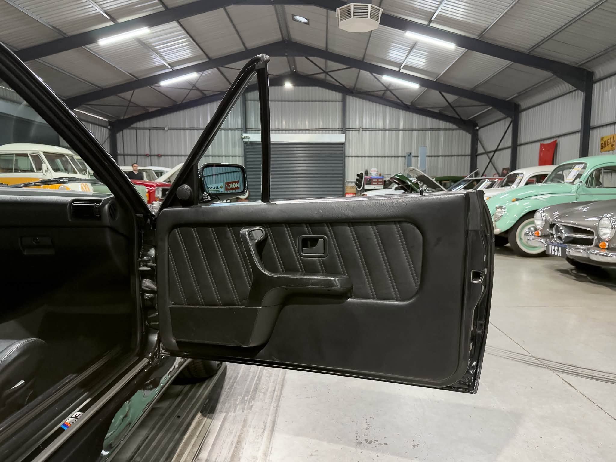 Inside a showroom/garage, the open passenger door of a black vintage car frames a row of classic cars under a metal warehouse ceiling.