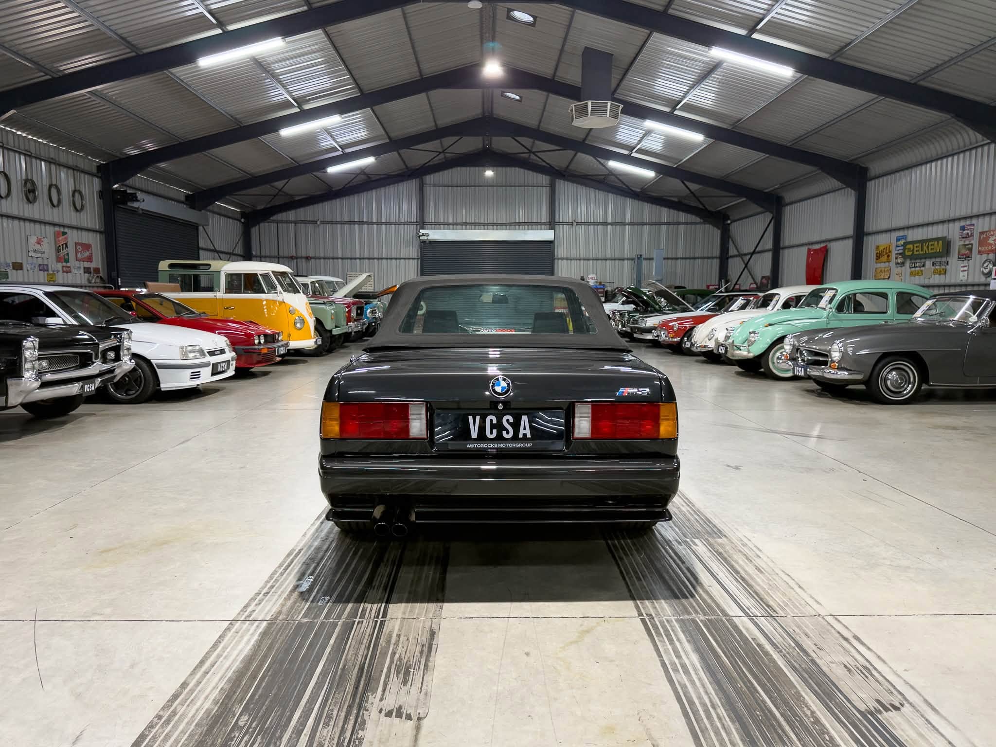 Interior of a large warehouse filled with vintage cars lined up along both sides, viewed from the rear of a black BMW M5 at center.