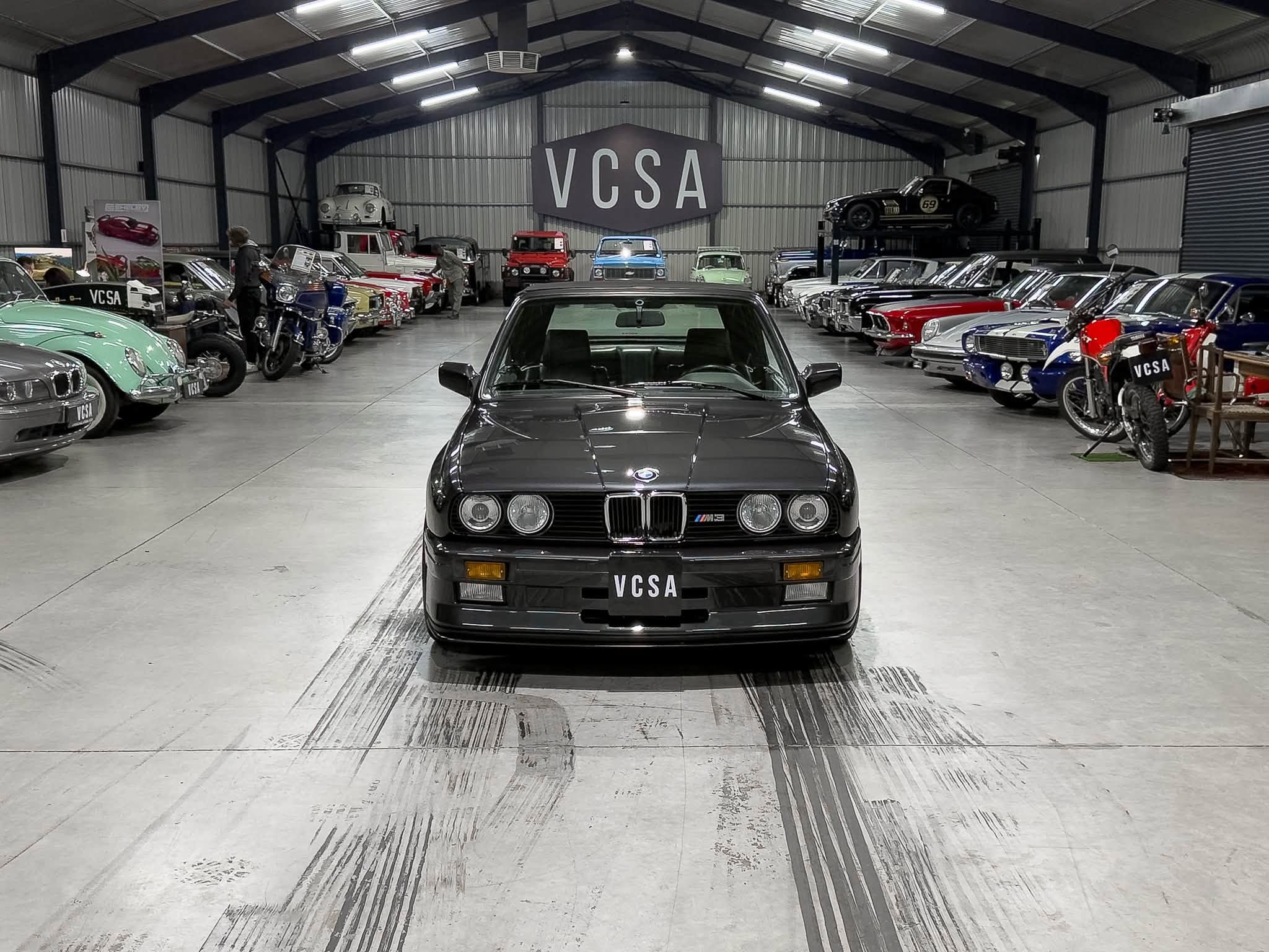 Inside a bright showroom filled with rows of vintage cars on both sides and a black BMW in the foreground with a VCSA plate label on the bumper, center stage.