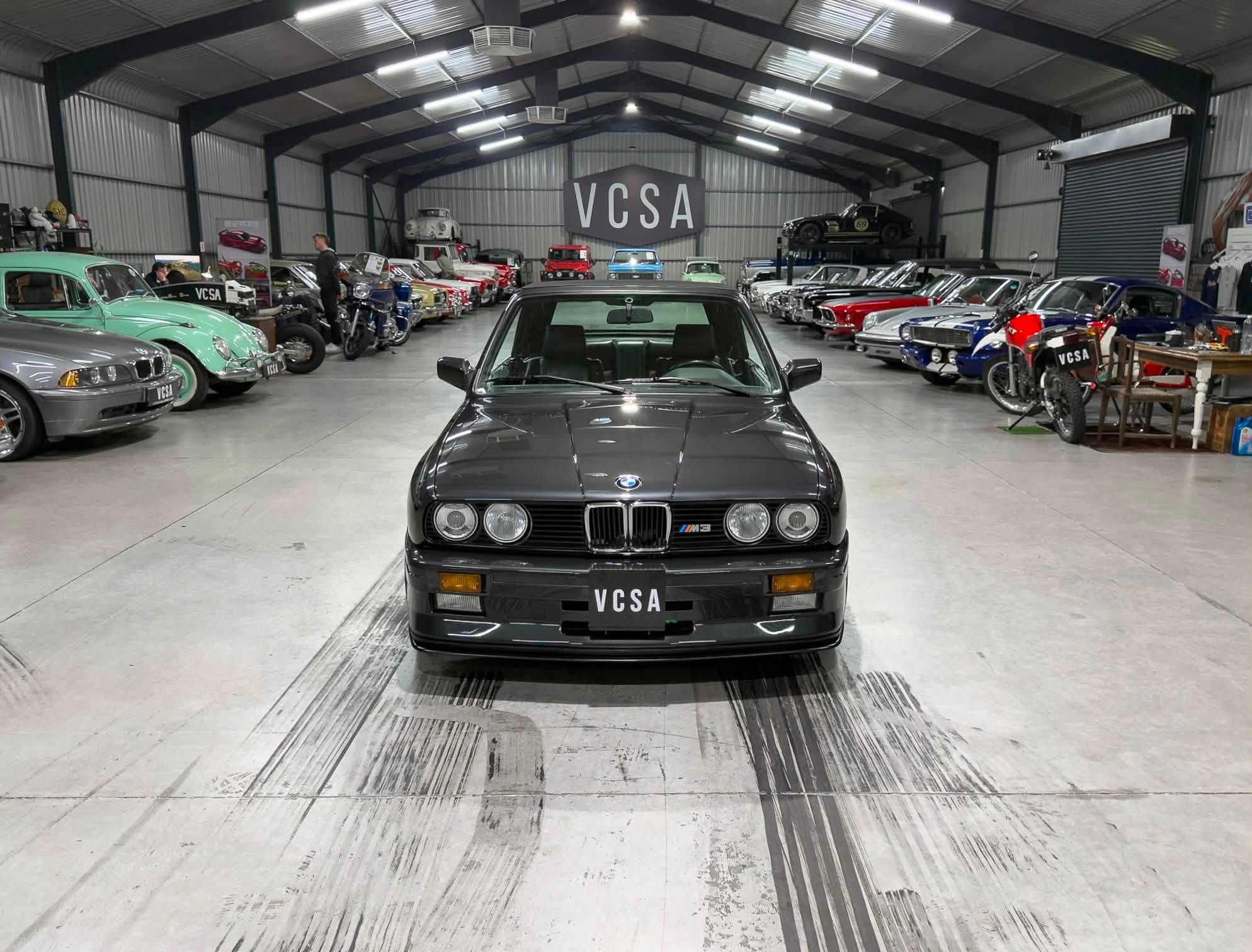 Inside a large showroom/garage filled with vintage and modern cars lined up on both sides, with a dark BMW M3 centered in the foreground and a VCSA sign in the back.