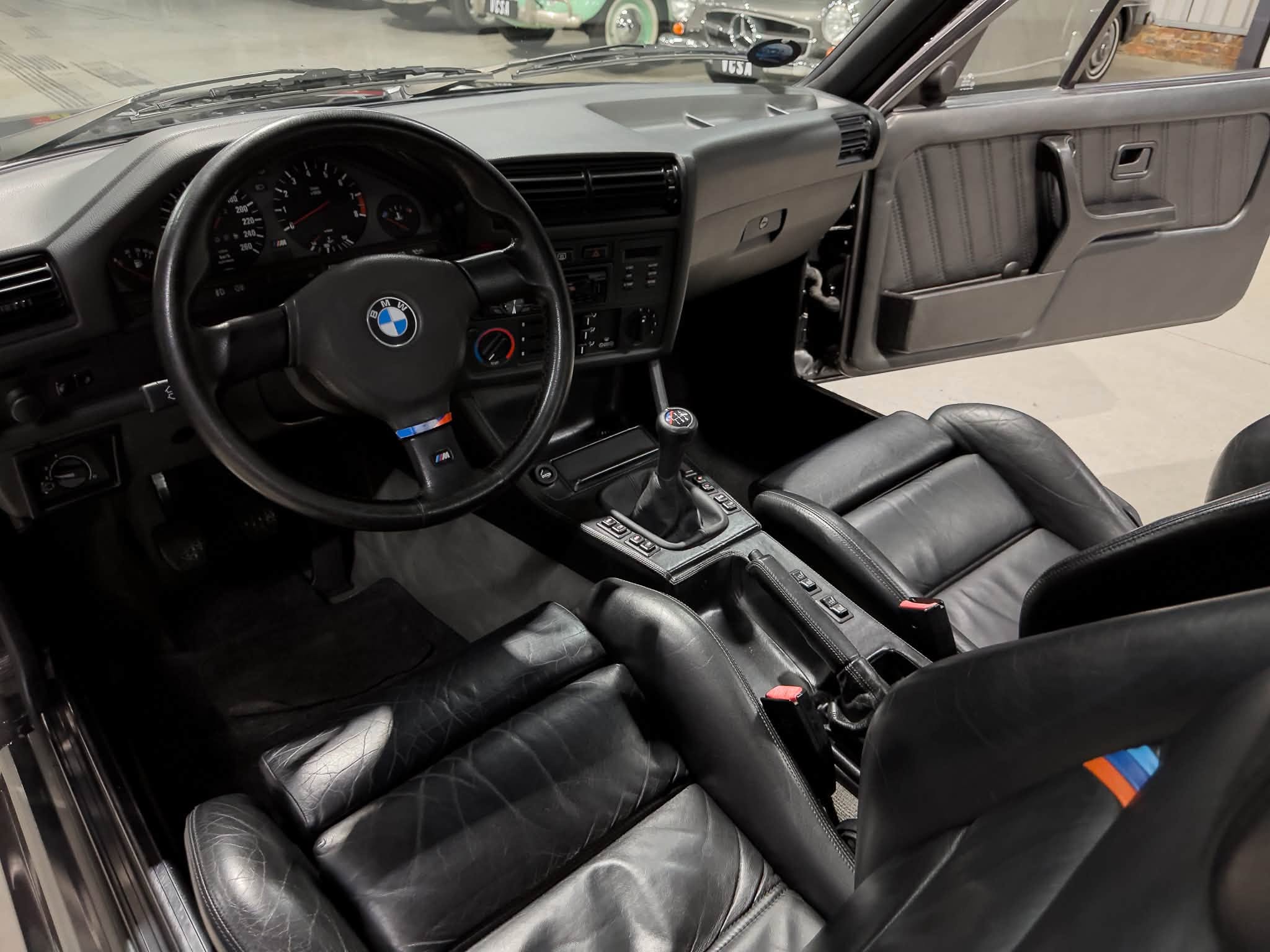 Interior view of a classic BMW car: black leather seats, steering wheel, center console, and manual gear shifter in a showroom setting with other cars in the background