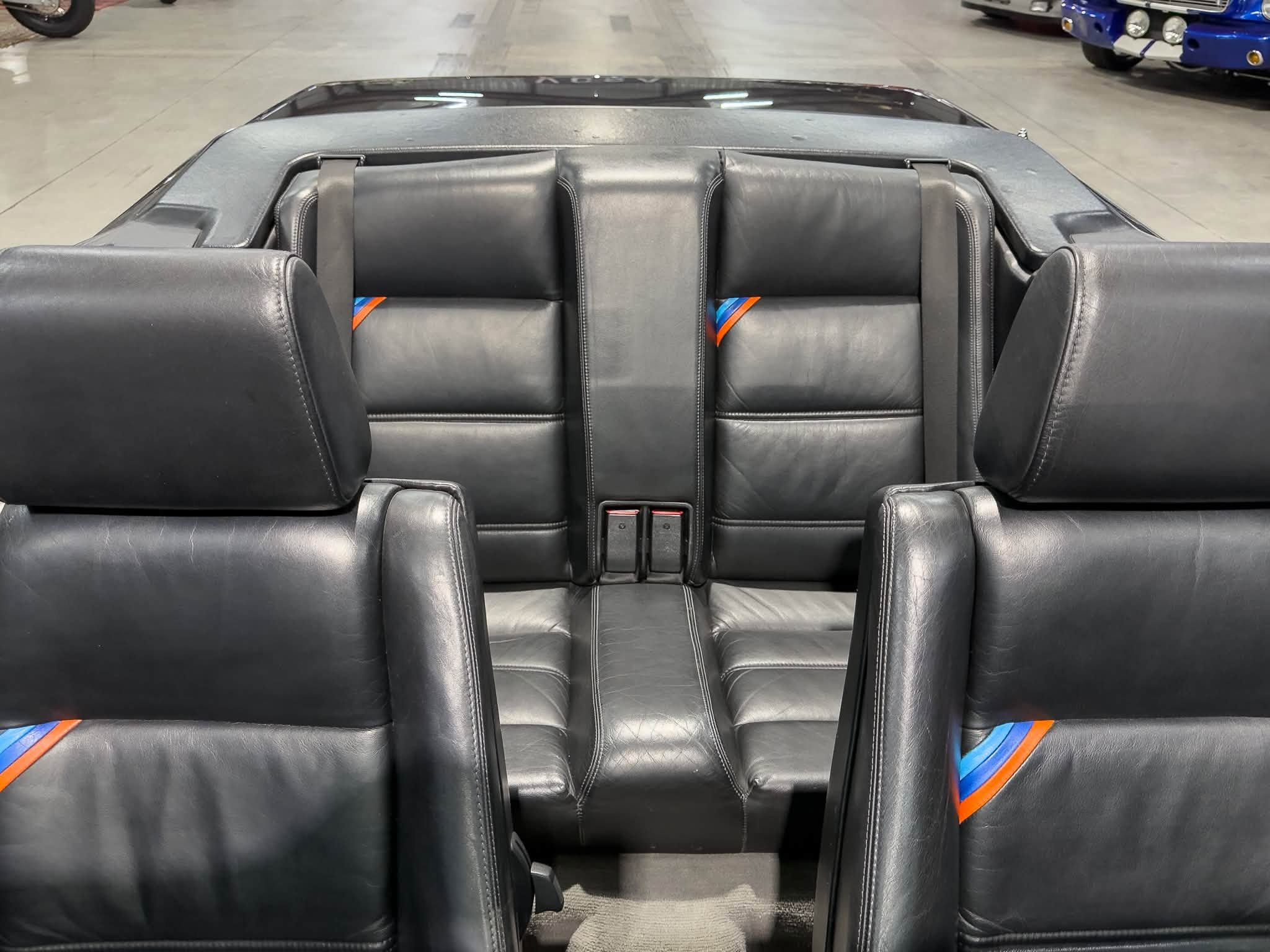 Rear seats of a black leather convertible with orange and blue seat belt accents in a showroom.