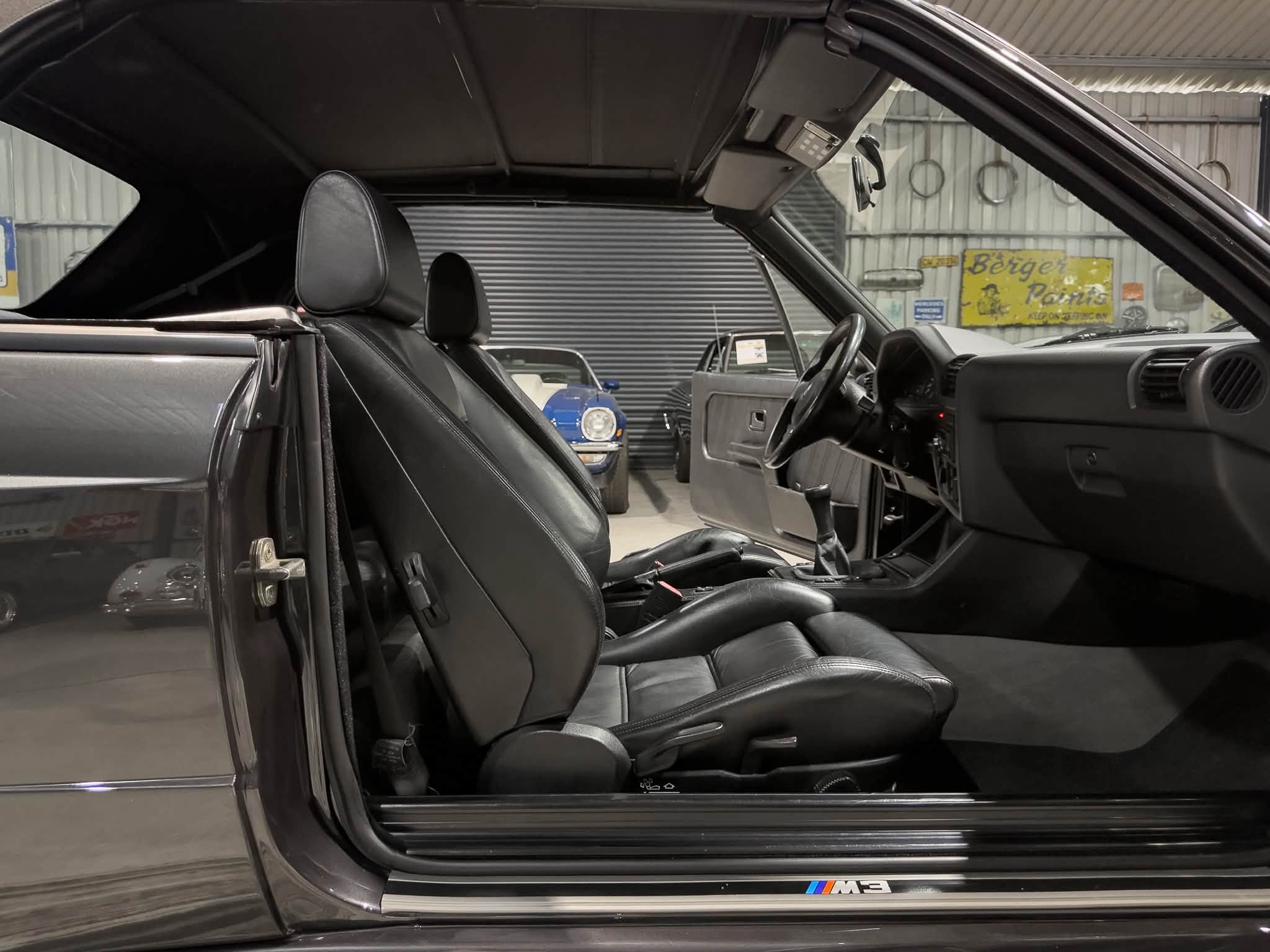 Open car door revealing black leather interior of a BMW M3, showing front seats and dashboard in a showroom setting.","Side open door of a BMW M3 with black leather seats and steering wheel visible; car badge on sill.","Interior view of a BMW M3 coupe with driver-side door open, highlighting the black leather seats and dashboard.","BMW M3 interior shot: open door, visible steering wheel, center console, and signature M stripes on the door sill.