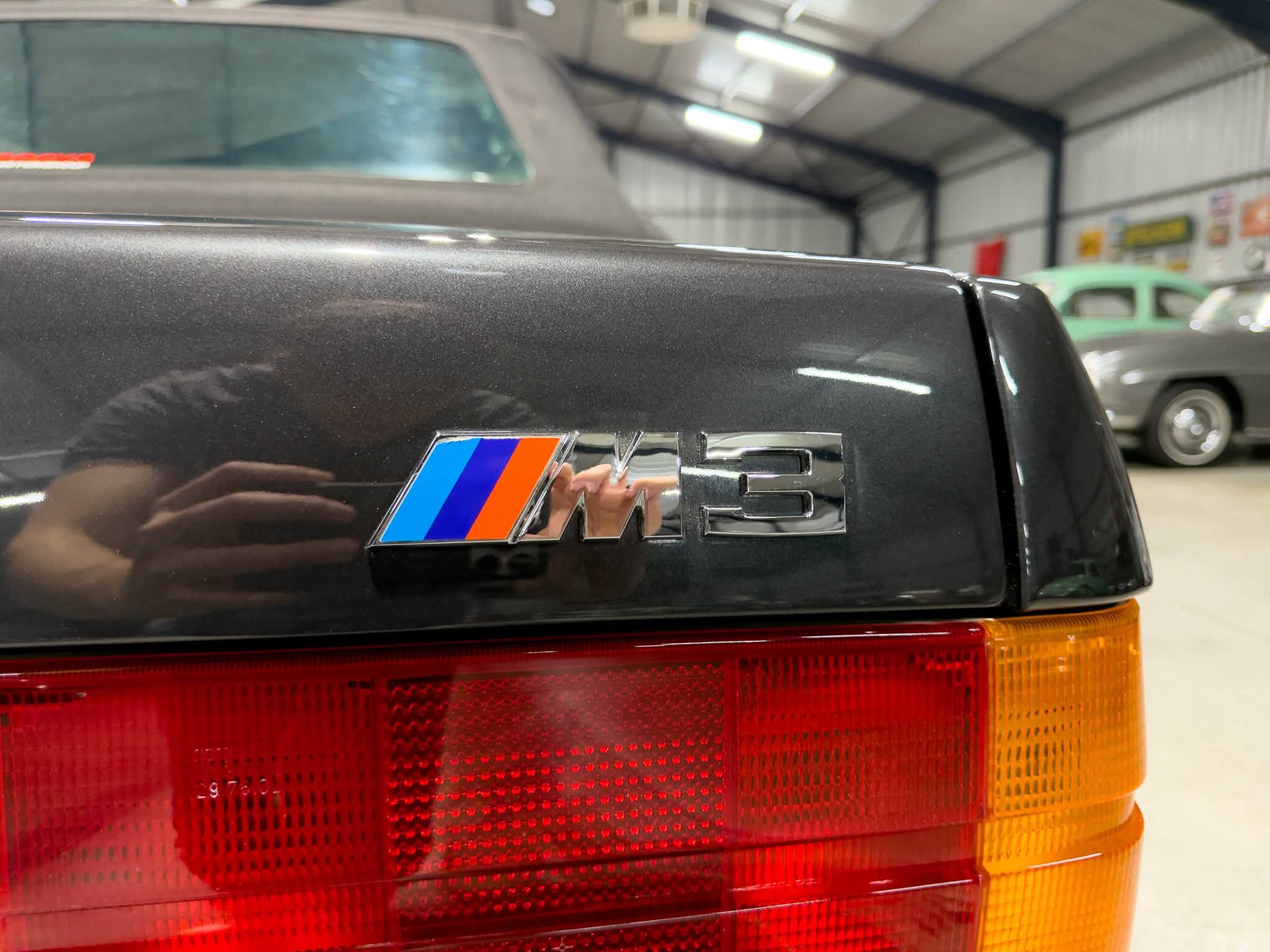 Close-up of the BMW M badge on a dark car trunk in a showroom, with red taillights below.