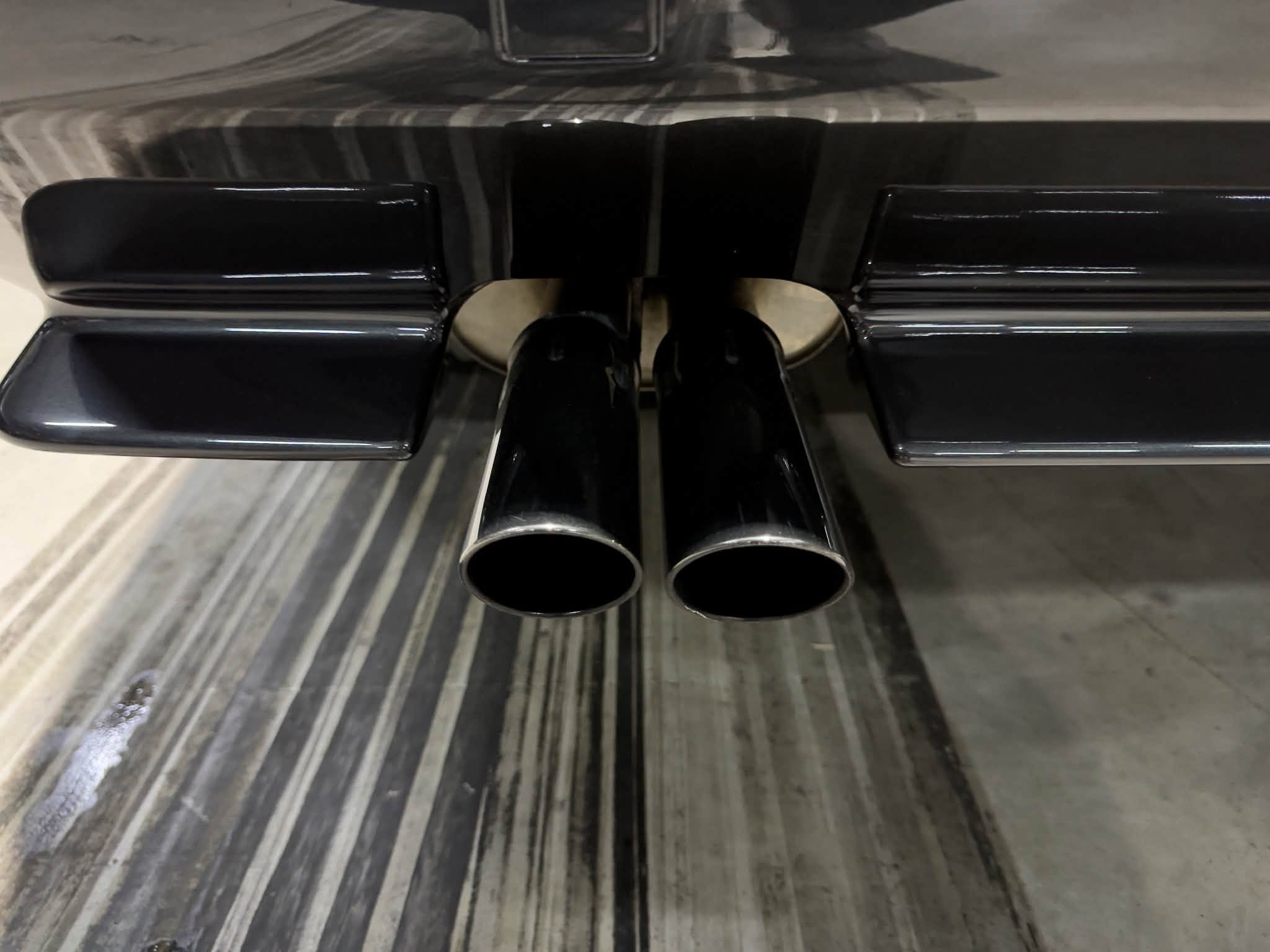 Dual black exhaust tips protruding from the rear of a glossy black vehicle, viewed from behind on a striped floor.