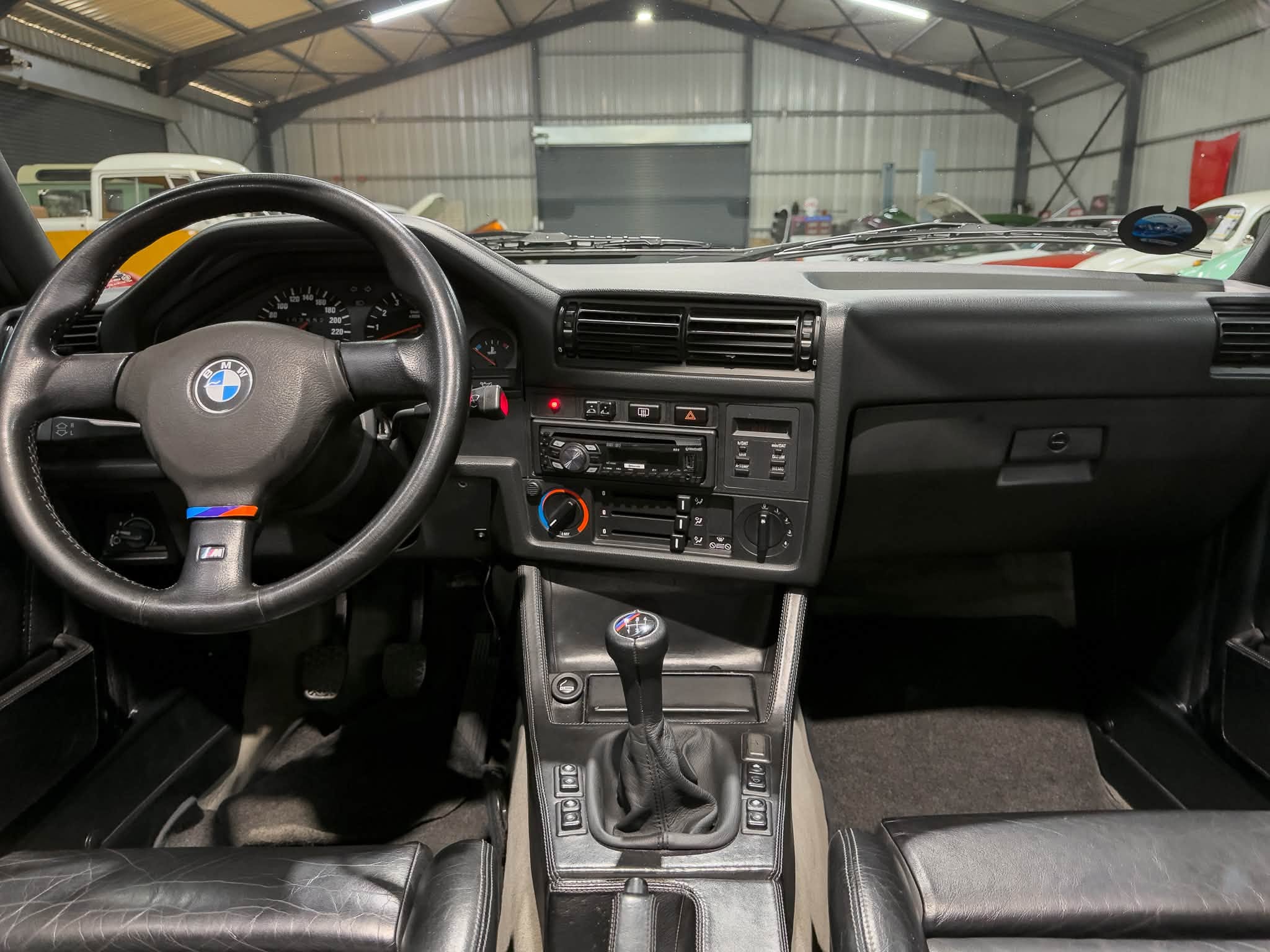 View from the driver's seat of a black BMW showing steering wheel, dashboard, center console, and gear shifter inside a garage with vintage cars outside.