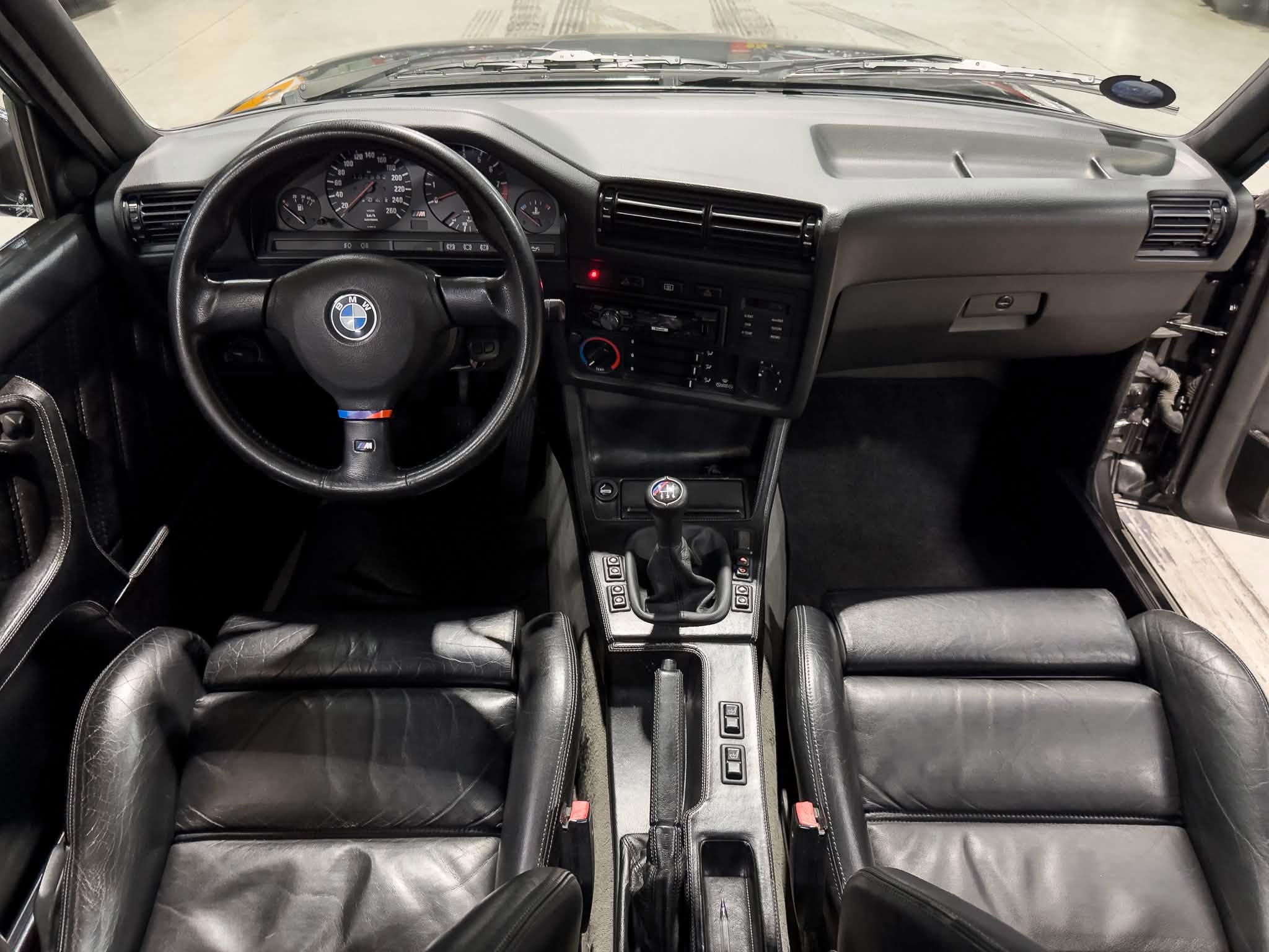 Left-hand-drive BMW interior with leather seats, steering wheel, and central console showing climate controls and gear shifter viewed from front seats